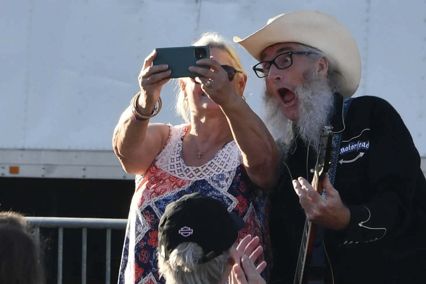 A woman takes a selfie with a man playing guitar, both appear excited. The man, wearing a cowboy hat and glasses, has a surprised expression, while the woman is partially obscured by her phone. There is a child wearing a black cap in front of them.