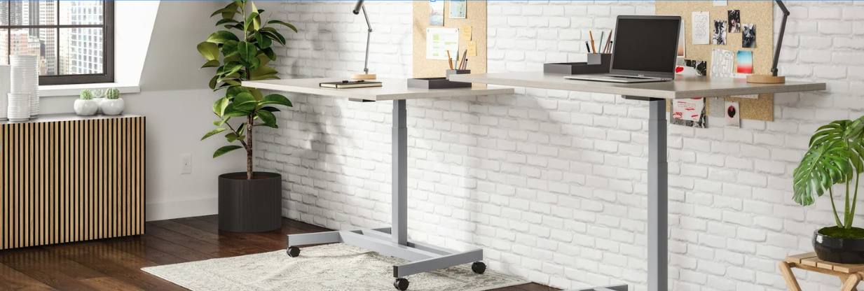Modern standing desk in a bright home office with a white brick wall, potted plants, cork boards with notes, and a window with city views.