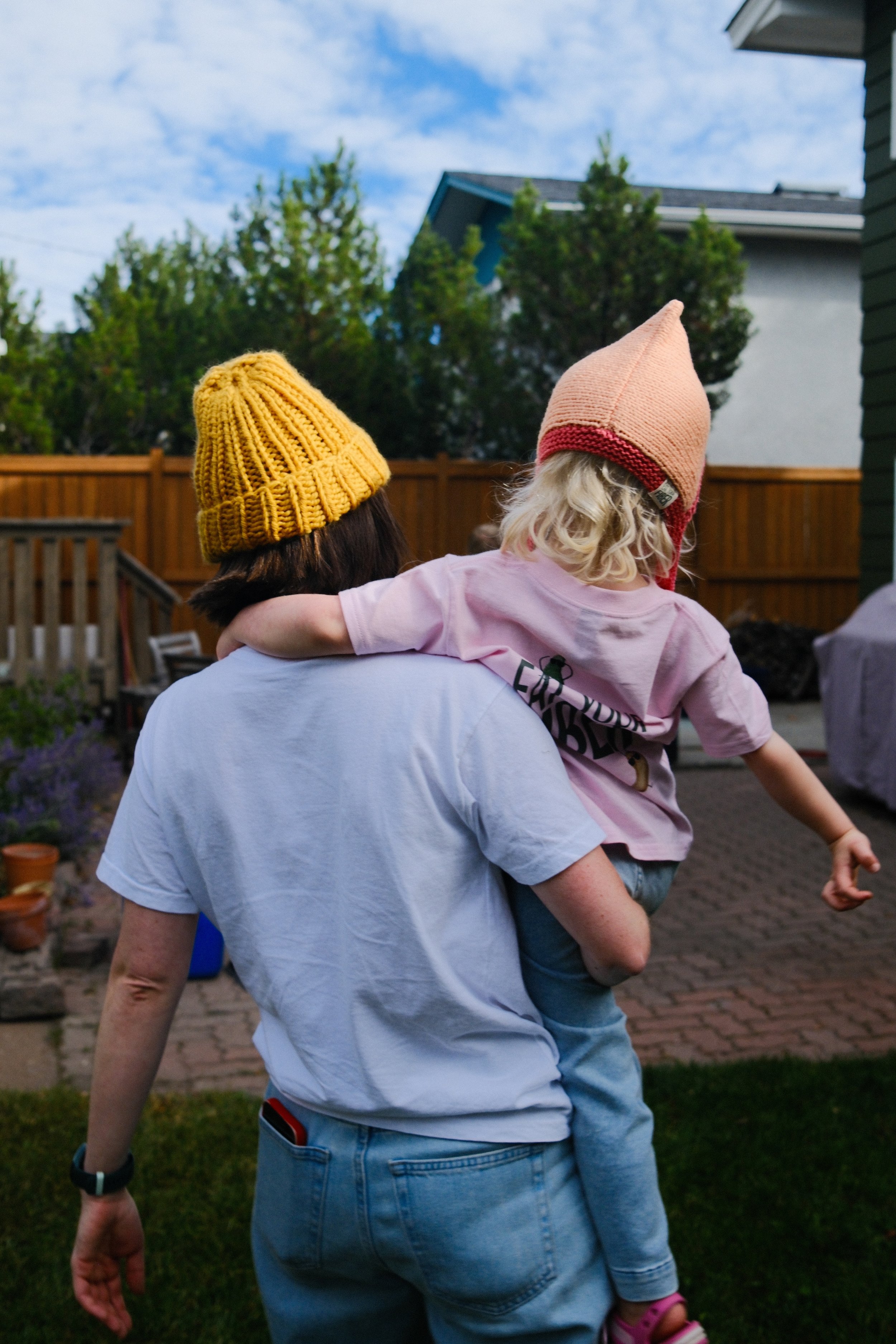 A person carrying a child outdoors in a backyard, both wearing colorful knit hats, with trees and a wooden fence in the background.