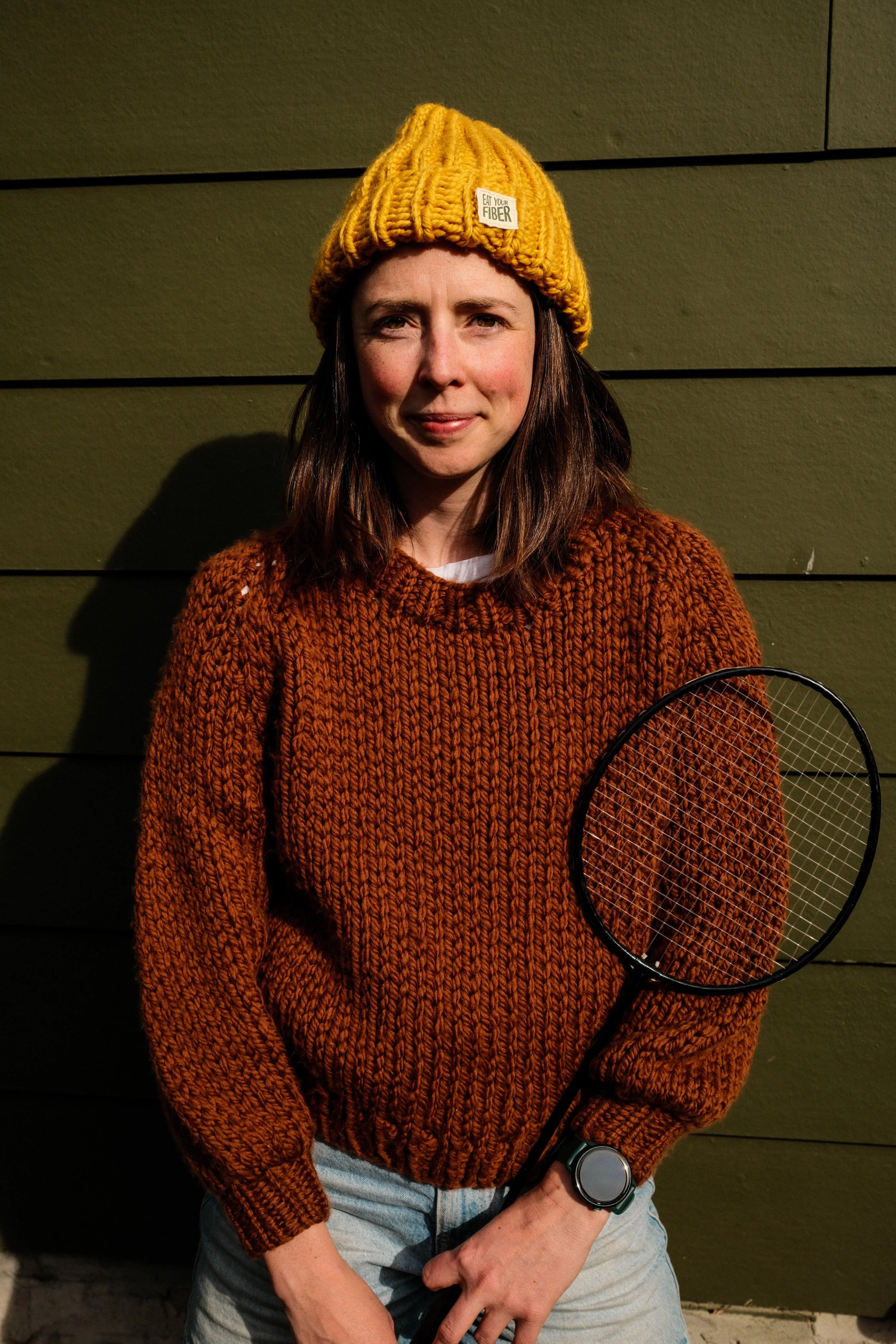 A woman wearing a yellow knit hat and a brown chunky knit sweater, holding a badminton racket, standing outdoors against a green wooden wall.