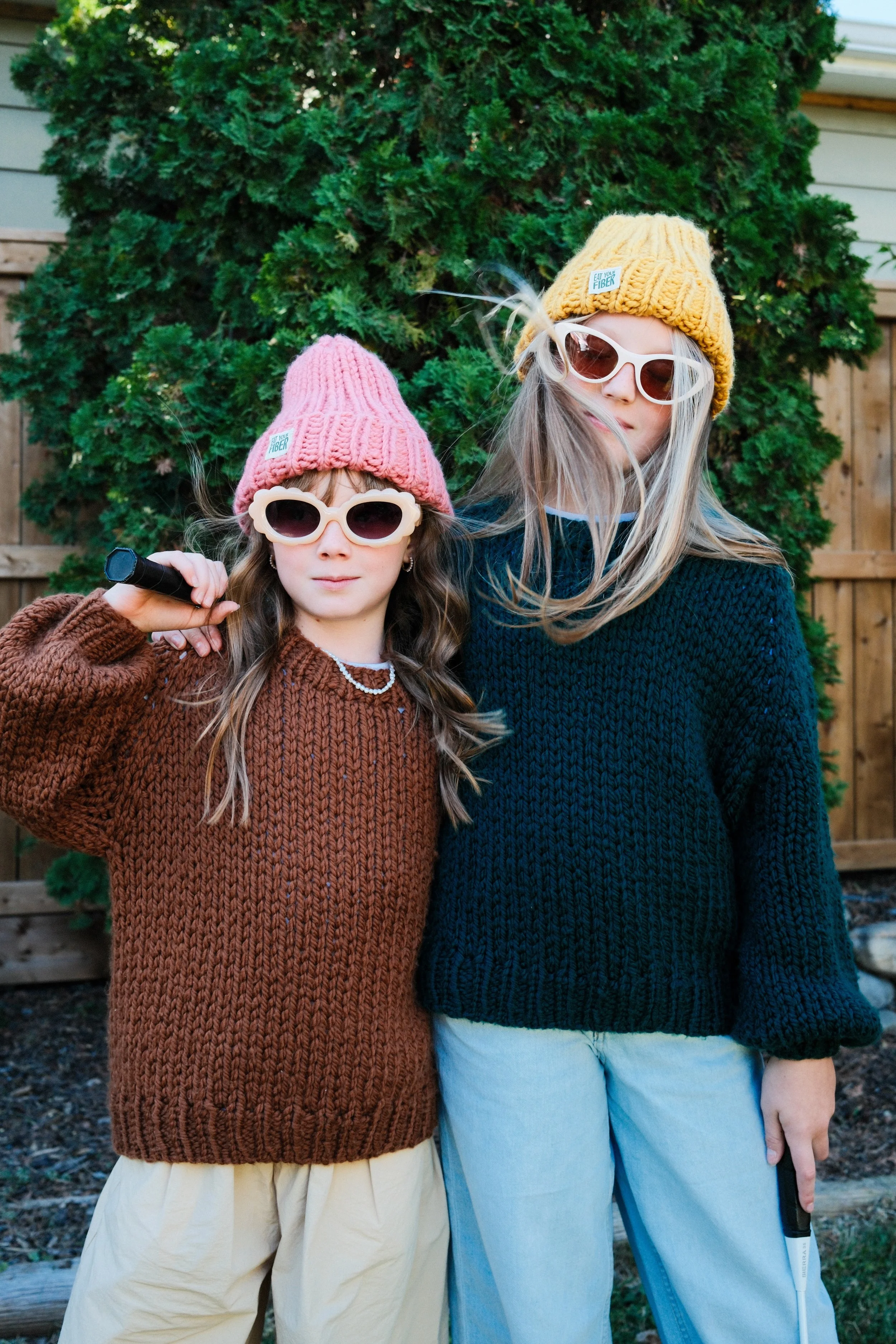 Two girls with sunglasses and knit hats standing outdoors in front of a green bush and wooden fence.