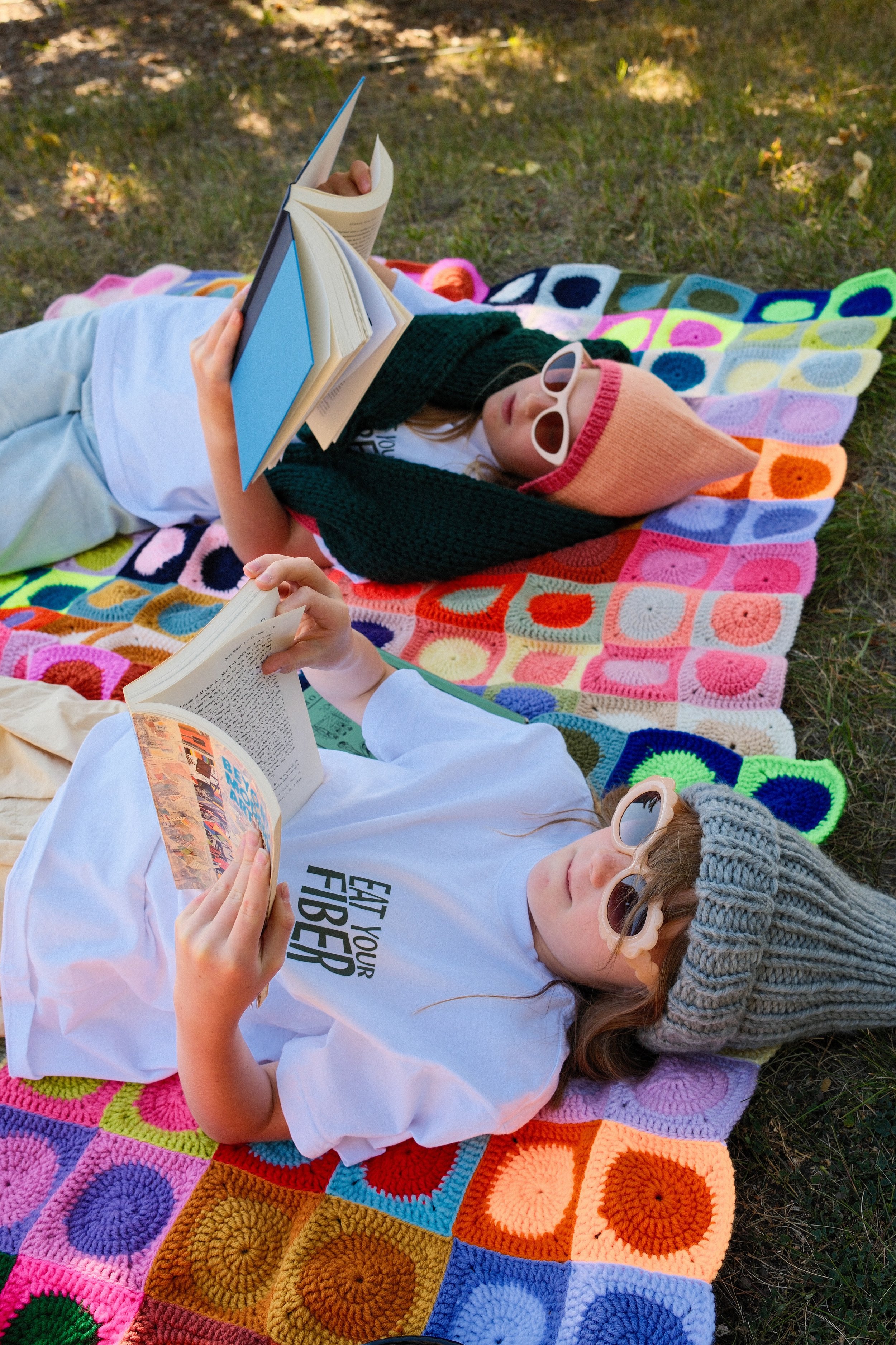 Two girls lie on colorful crocheted blankets outdoors, reading books. One girl wears a gray knit beanie and pink sunglasses, the other wears a pink knit bonnet and white sunglasses.
