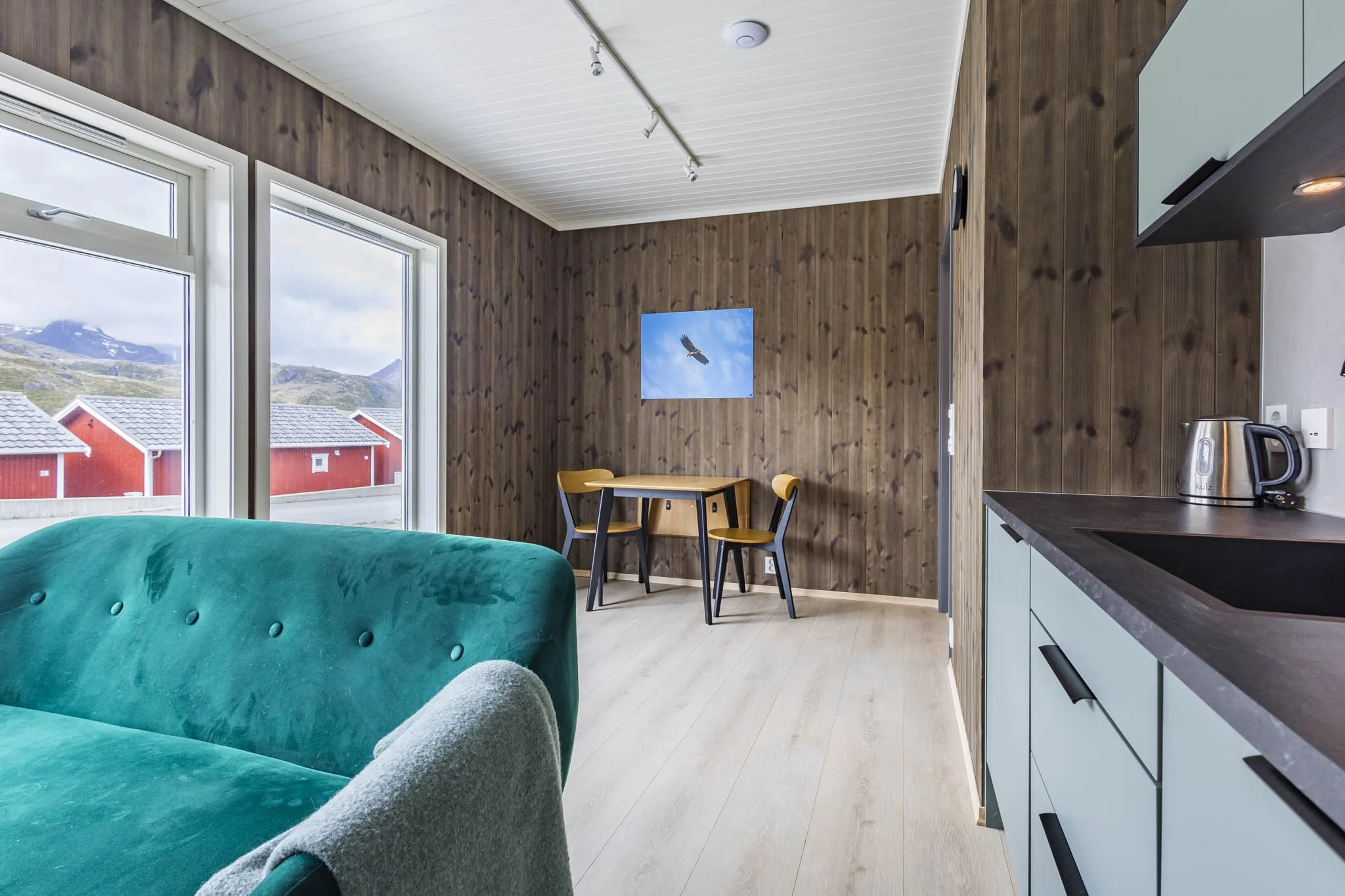 Interior of a modern room with wood-paneled walls, large windows revealing mountains and red buildings outside, a teal sofa, a small dining table with two chairs, a wall art of a bird, and kitchen appliances including a kettle.