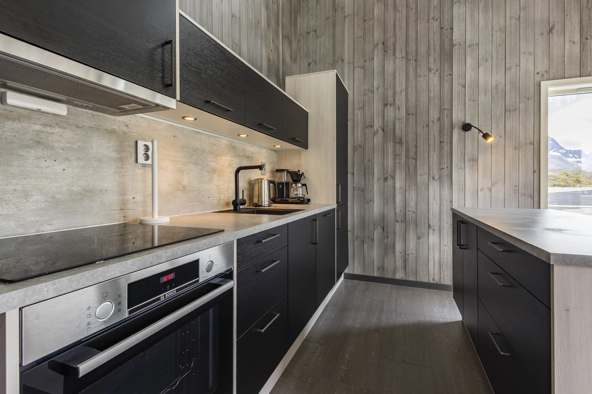Modern kitchen with black cabinets, a built-in oven, a sink, coffee maker, kettle, and a window with landscape view, wooden wall paneling, and gray floor.