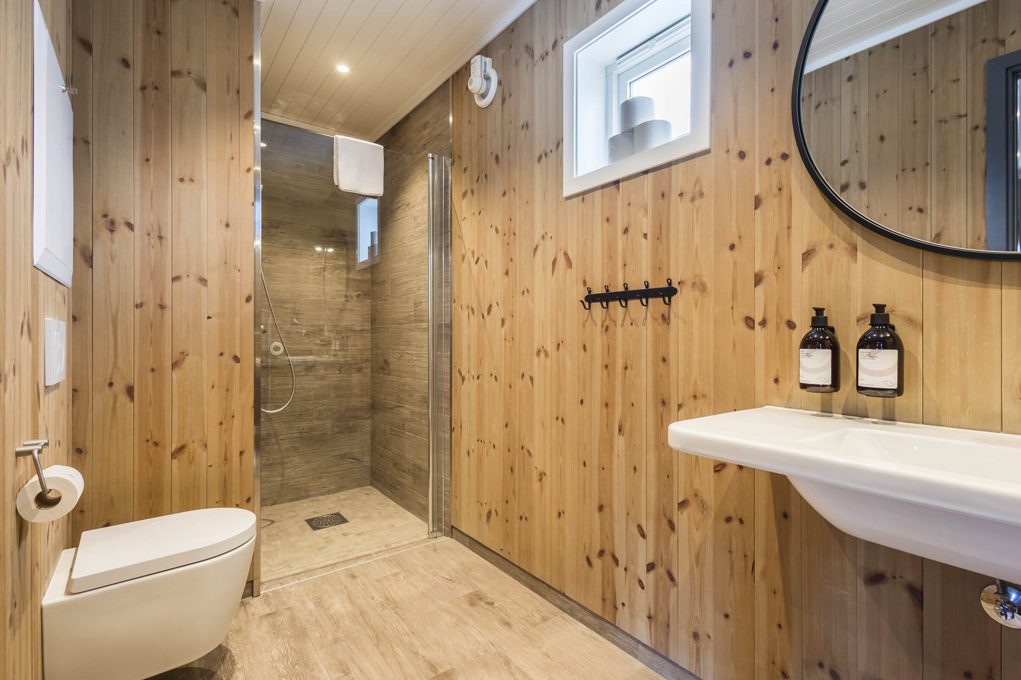 A bathroom with wood-paneled walls, a glass-enclosed shower, a modern toilet, and a small white sink with two black soap dispensers. There is a small window with three white candles on the ledge and a black towel hook on the wall.