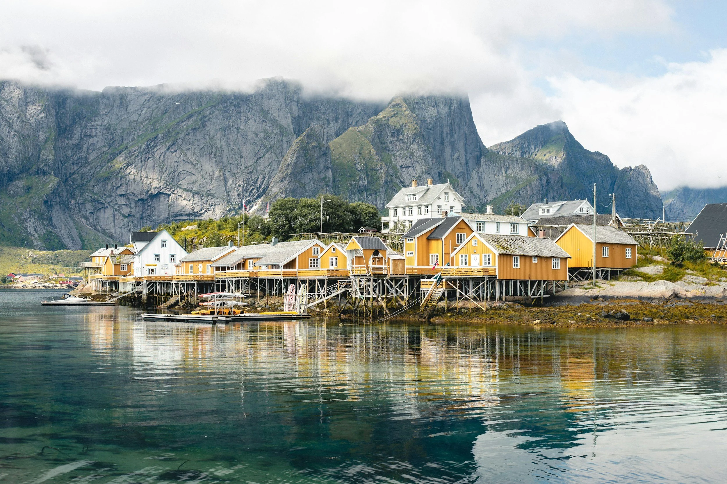Colorful houses on stilts along a calm waterfront with mountainous cliffs partly covered by clouds in the background.