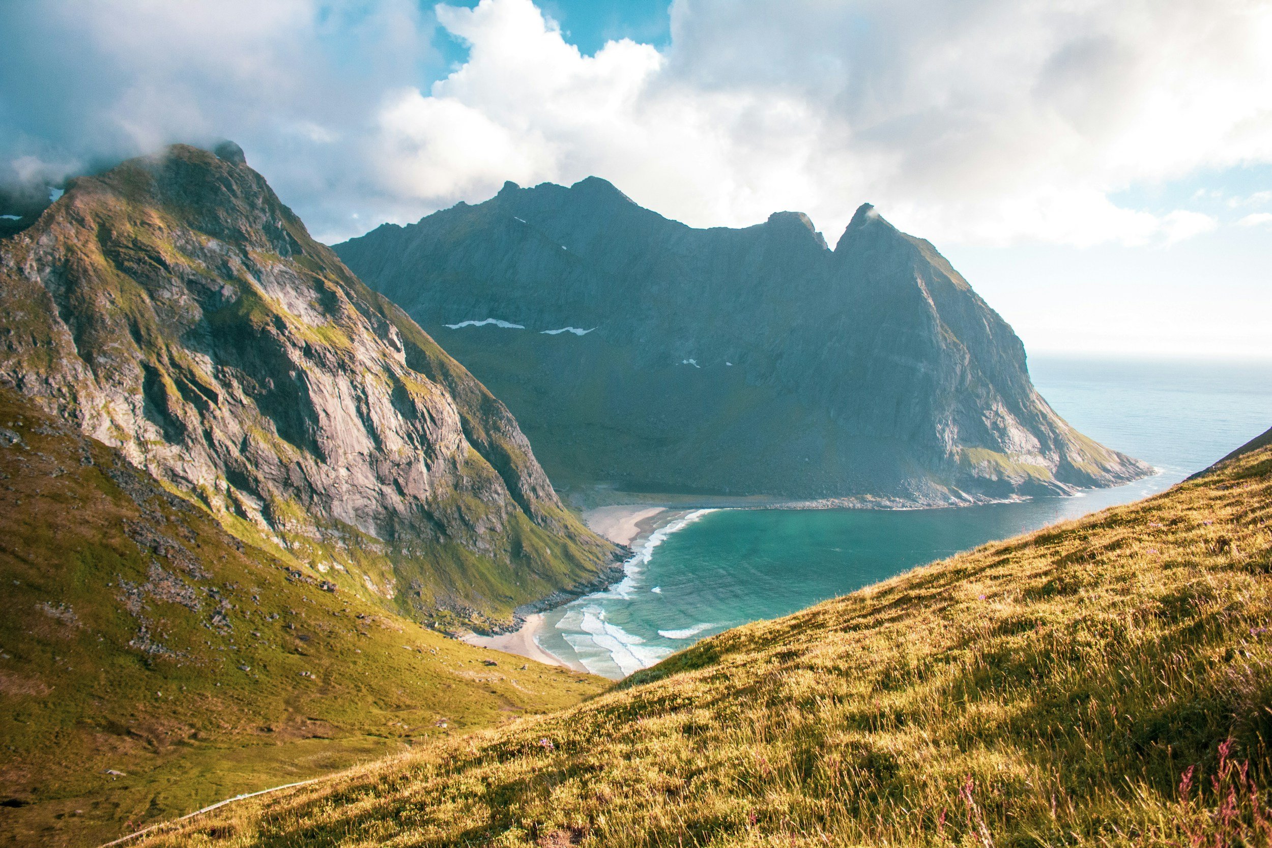 Coastal mountains with rocky, grassy slopes and a beach in a cove with turquoise water.