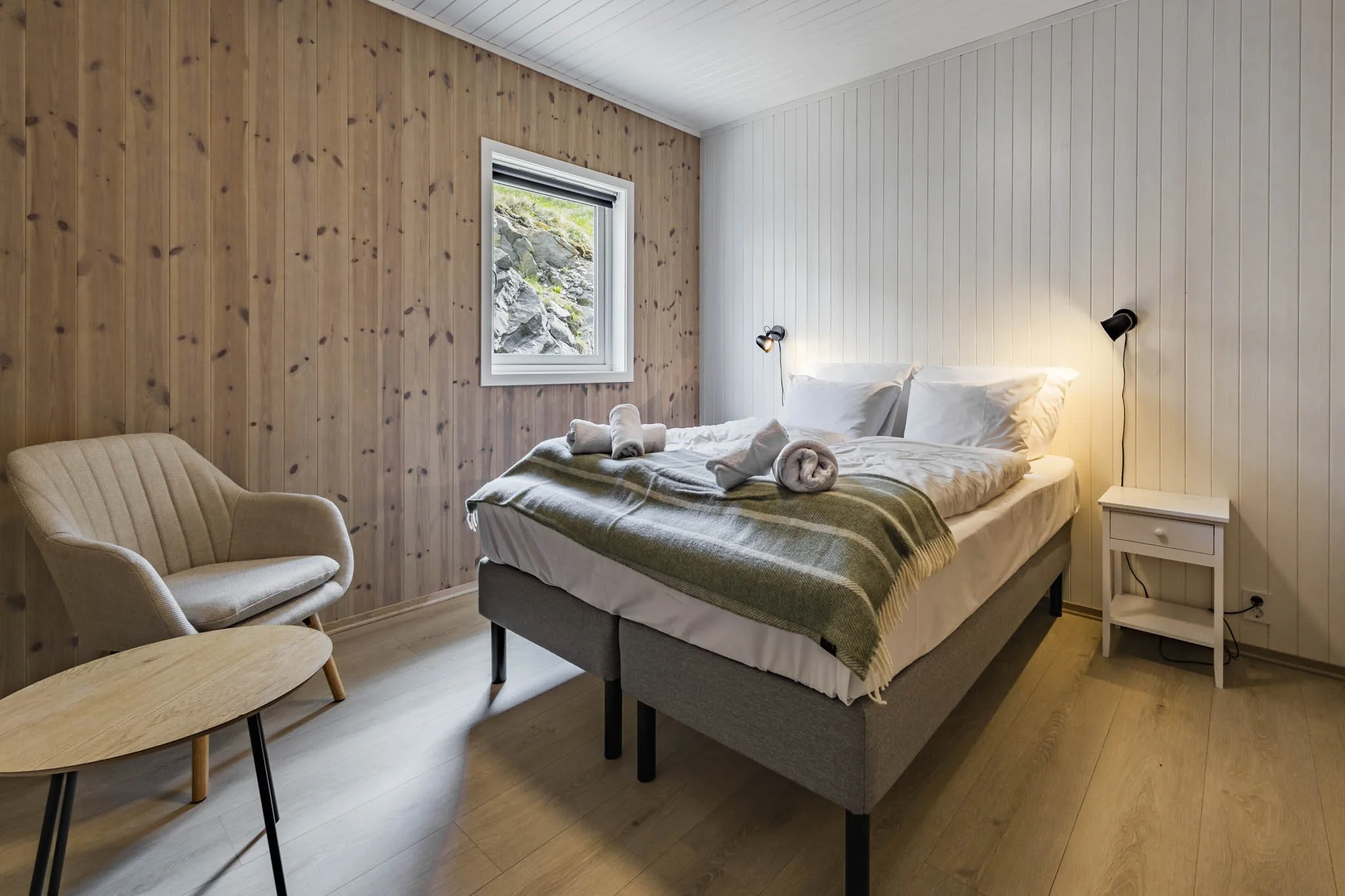 A bedroom with a wooden wall, a window showing rocks outside, a bed with white bedding and a green striped blanket, a small white nightstand, a wall-mounted reading lamp, a beige armchair, and a small wooden table with black legs.