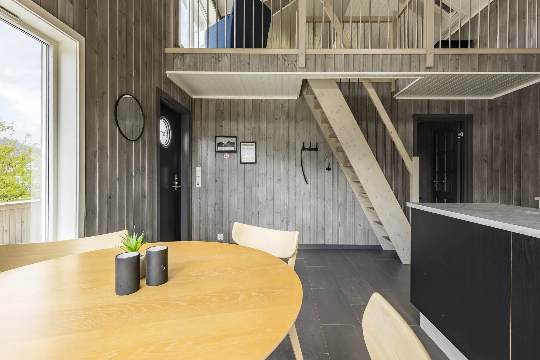 Interior of a house with wooden walls, a dining table with chairs, a staircase leading to a second floor, and decorations including wall art and a small window.