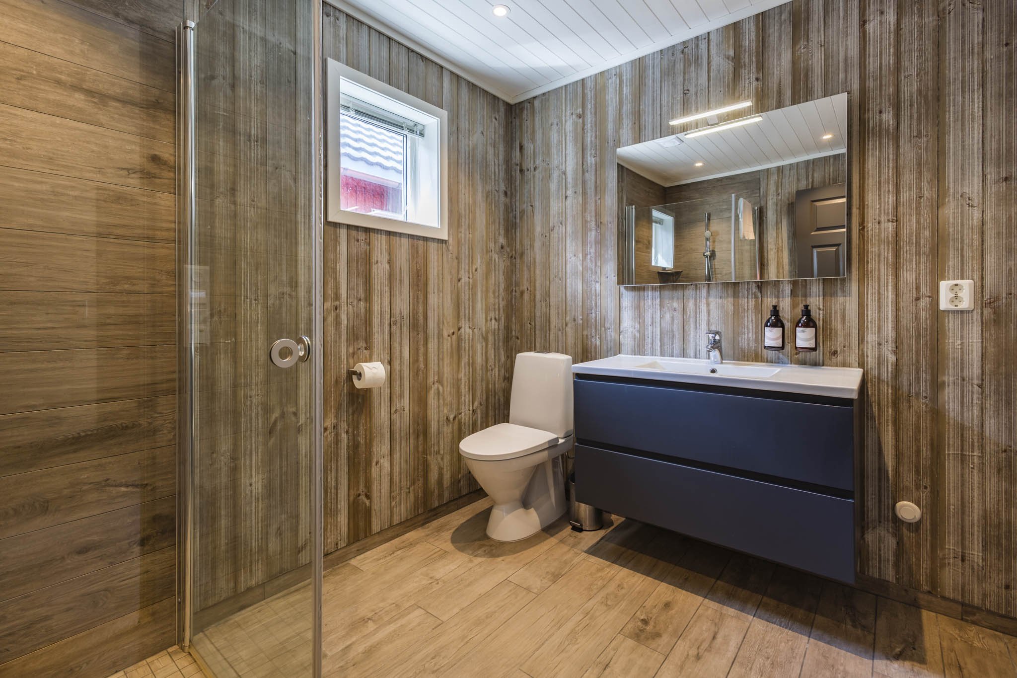 A bathroom with wooden wall panels and a wooden floor, a small window, a large mirror, a white toilet, a dark vanity with a sink, and two bottles of soap or lotion on the counter.
