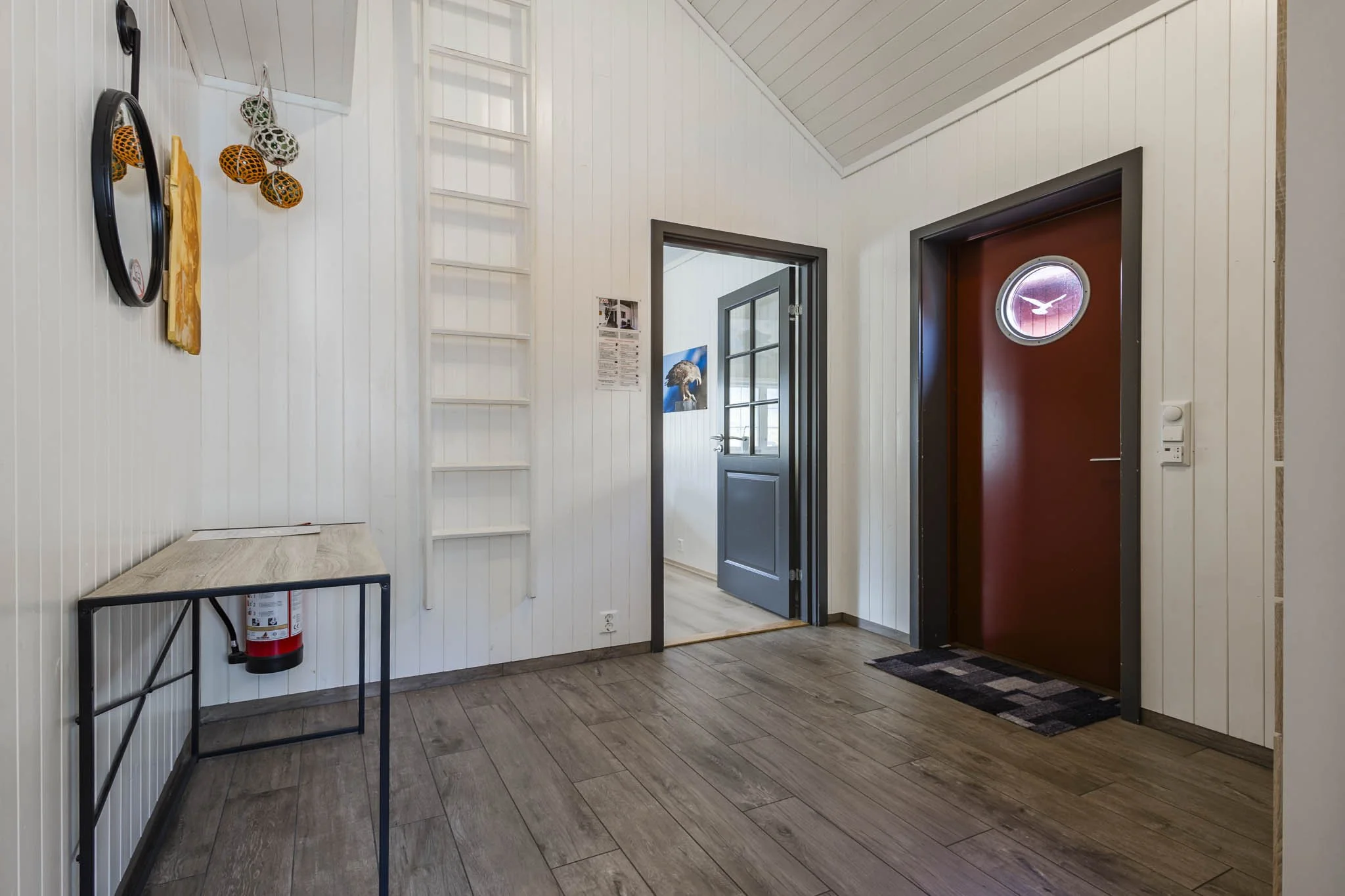 Interior of a house entryway with white paneled walls, a red door with a circular window featuring a bird silhouette, an open gray door leading to another room, a small black table, a fire extinguisher, and a decorative hanging with orange and white 