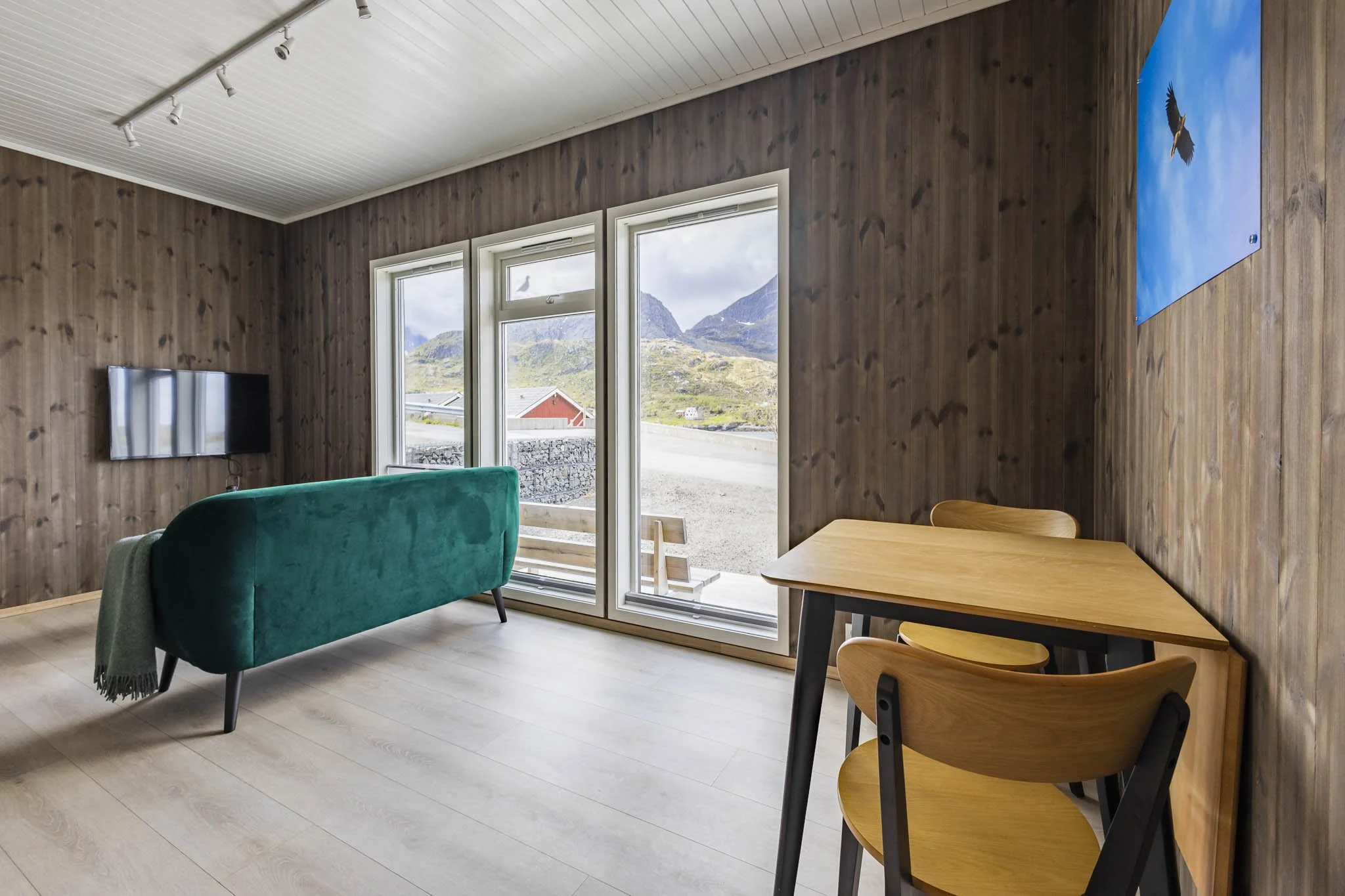 Living room with wooden walls, a teal sofa, a wooden dining table with chairs, a mounted flat-screen TV, and large windows showing mountains and a red building outside.