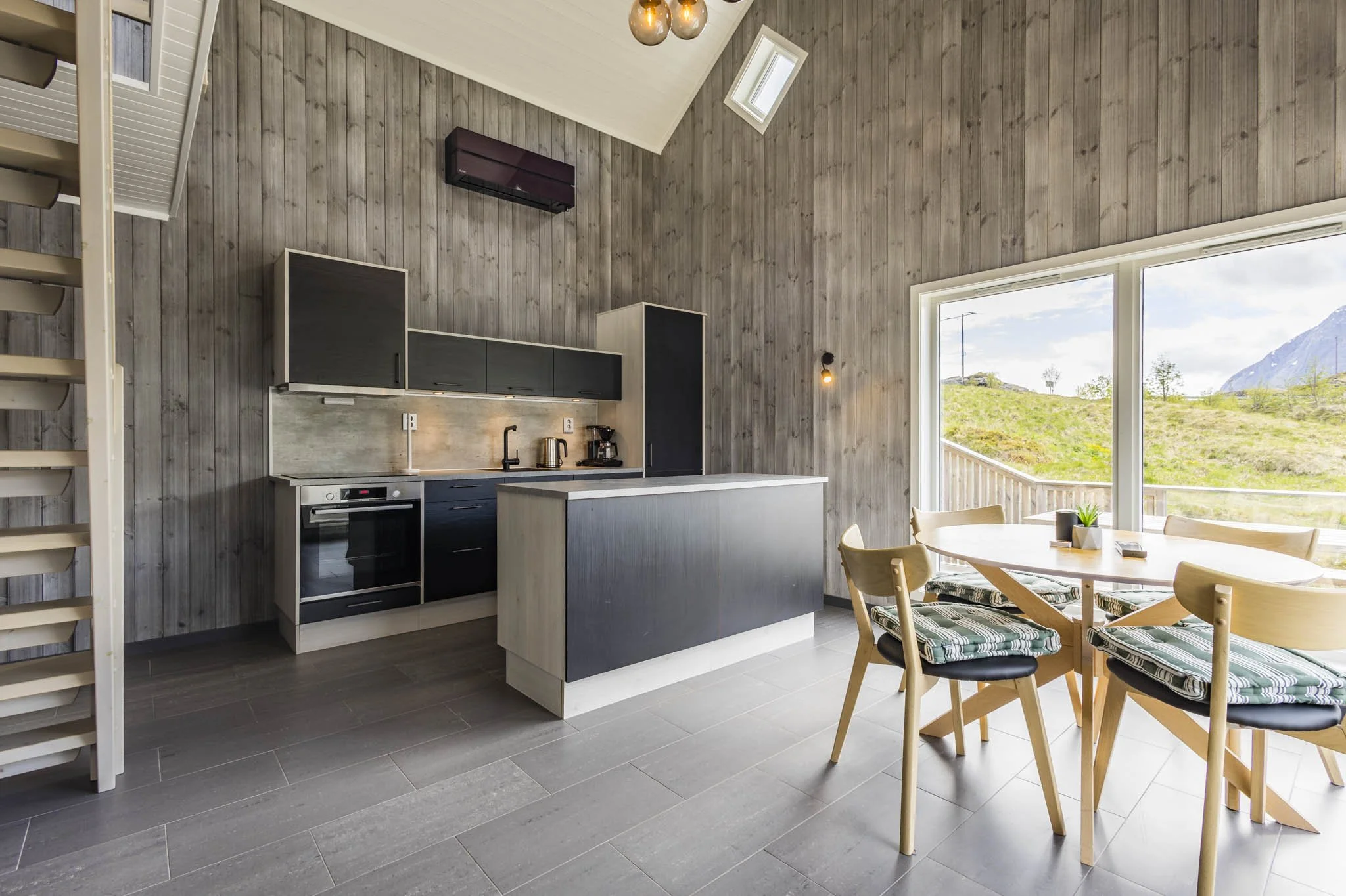 Modern kitchen and dining area with wood-paneled walls, large window showing a mountain and green landscape, round wooden table with four chairs, kitchen cabinets, stove, and small appliances, in a bright and airy space.
