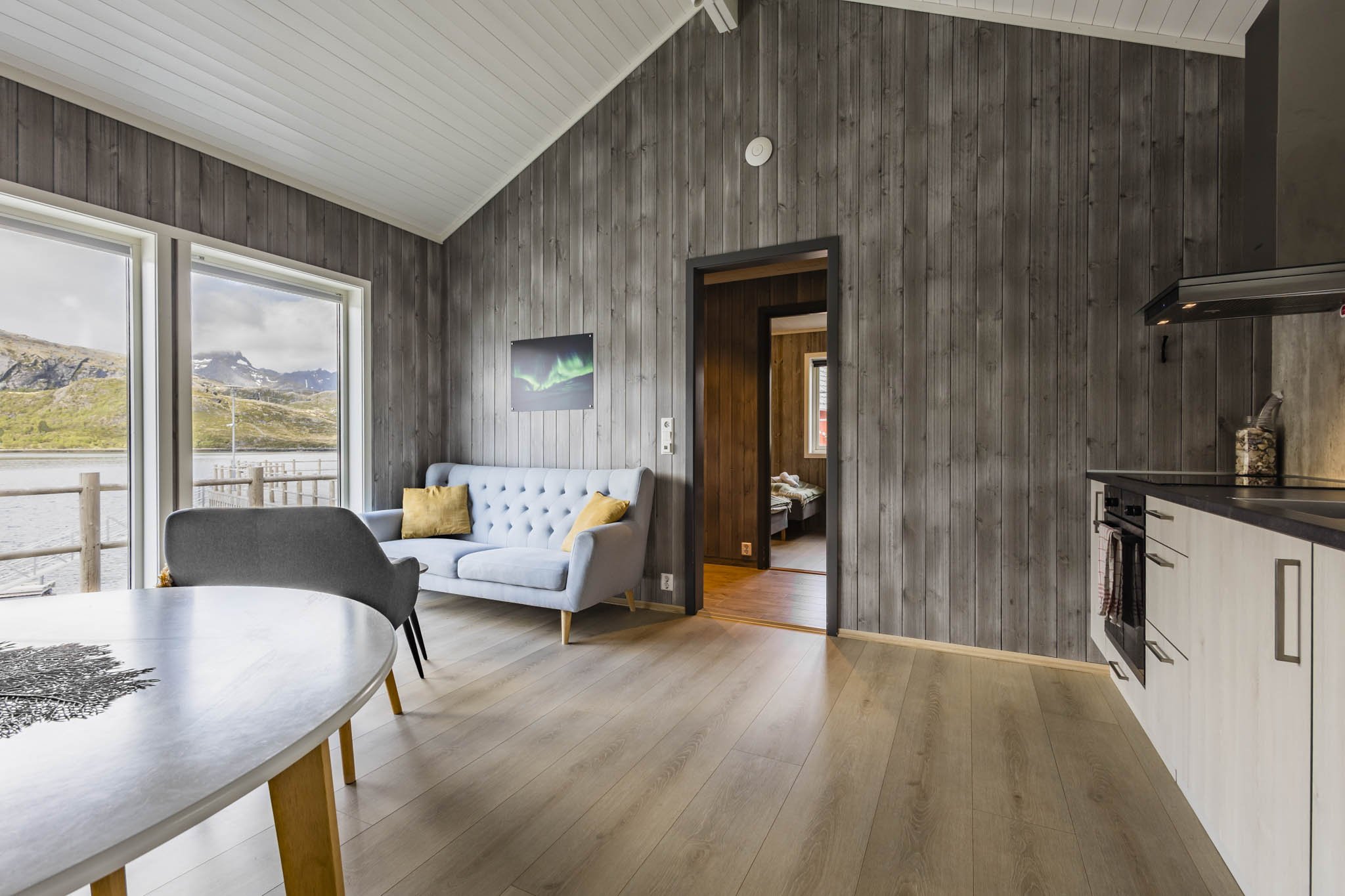 Interior view of a living room with gray wooden walls and a ceiling, large windows showing a mountain landscape, a white tufted sofa with yellow pillows, a white dining table, and a view into a small bedroom.