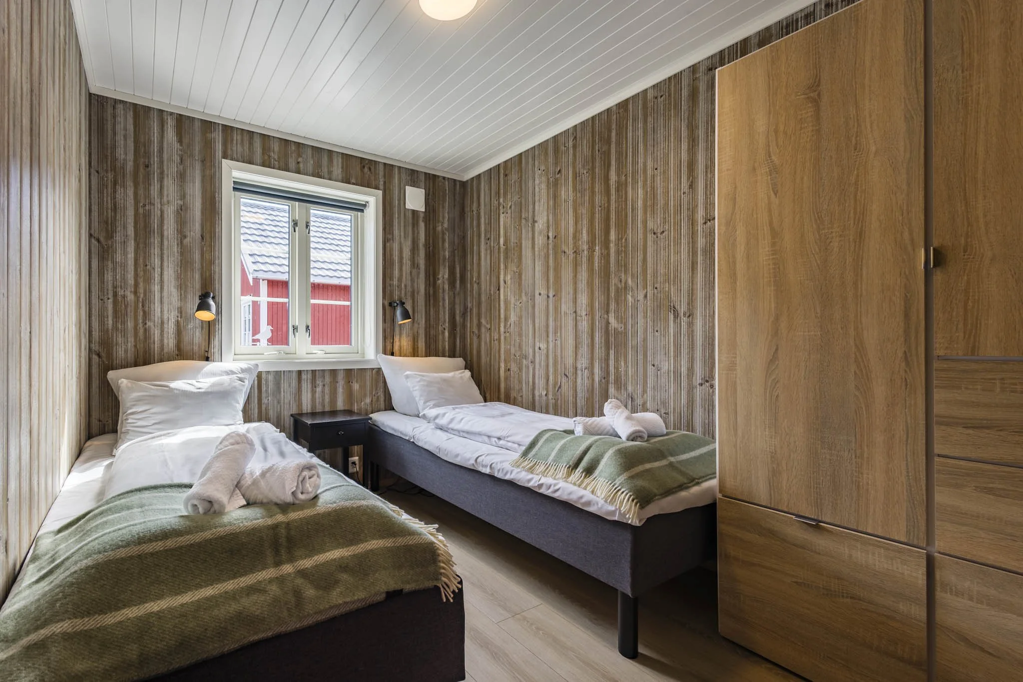 A cozy bedroom with wood-paneled walls, two single beds with white linens and green throws, a window showing a red building outside, and a wooden wardrobe on the right.
