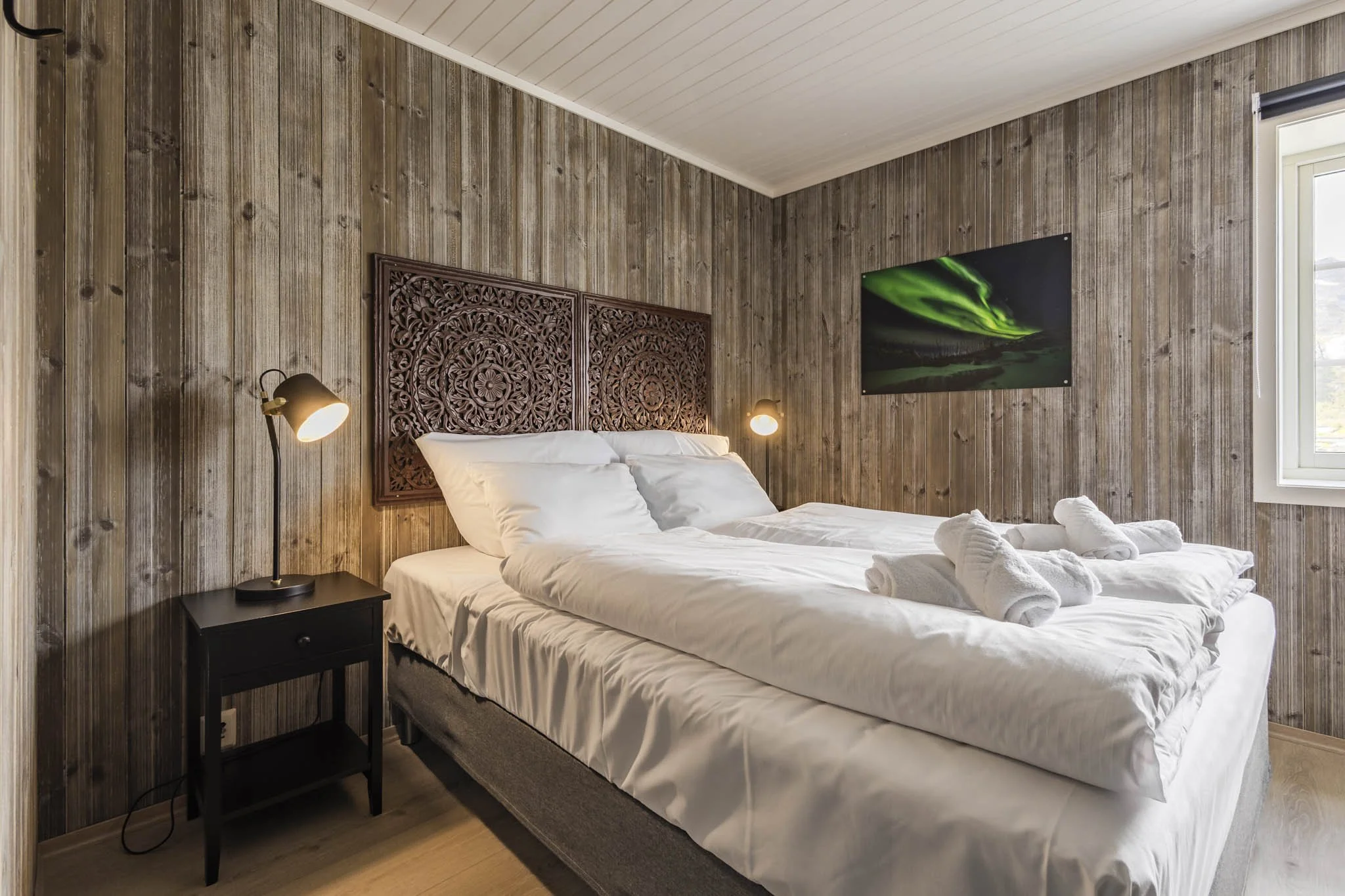 A cozy bedroom with wooden paneled walls and a sloped ceiling. There is a bed with white bedding and towels, a decorative carved headboard, a black nightstand with a lamp, and a wall-mounted TV displaying an image of the northern lights. A window wit