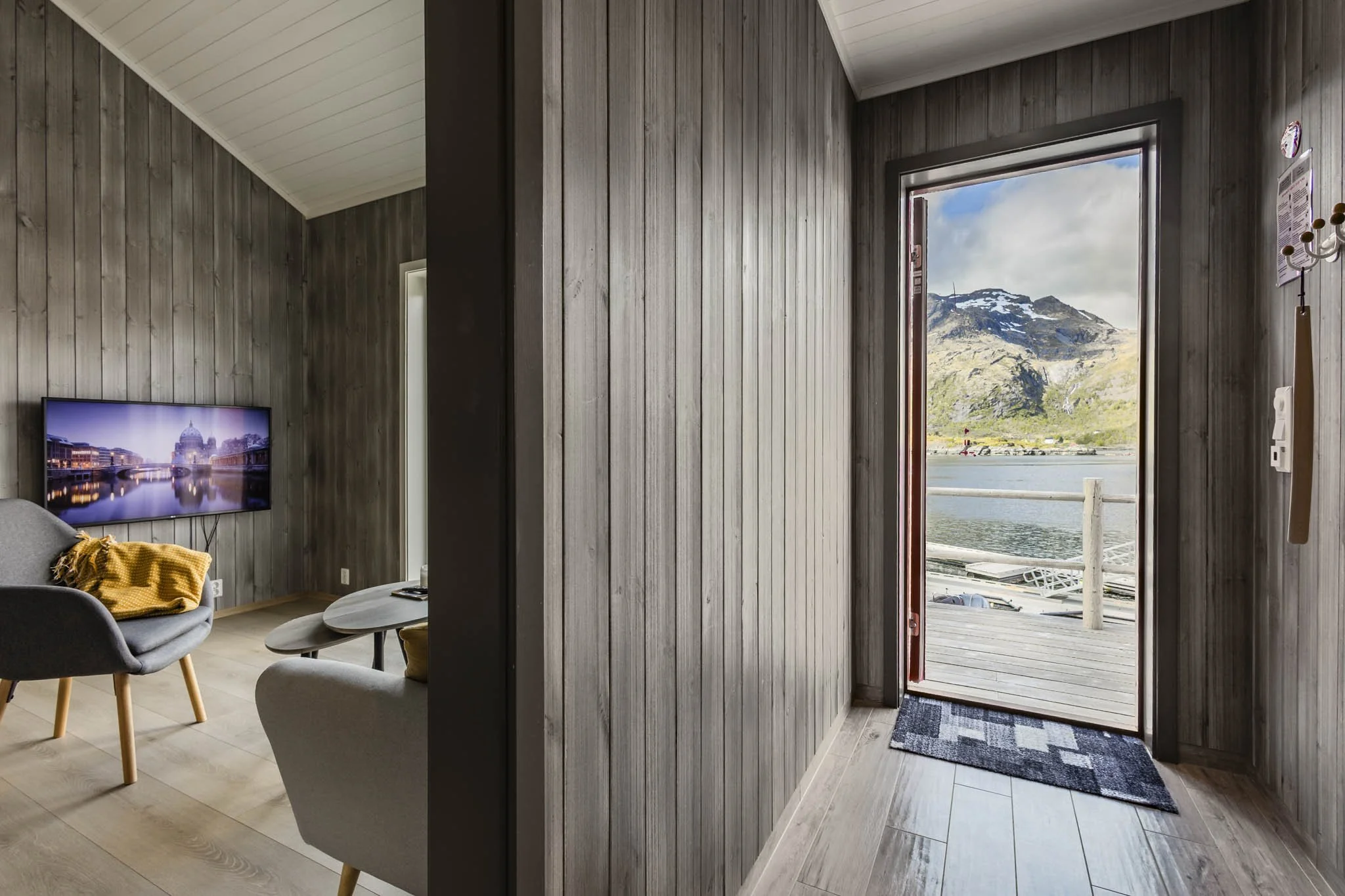 Living room with wood-paneled walls and a door leading to a deck, overlooking mountains and a body of water.