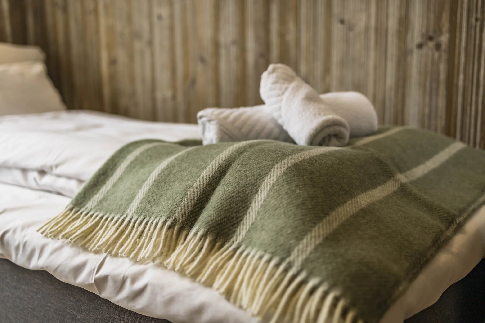A bed with a green and beige striped blanket, white pillows, and a folded towel on top of the blanket near the pillows. The bed is against a wooden-paneled wall.