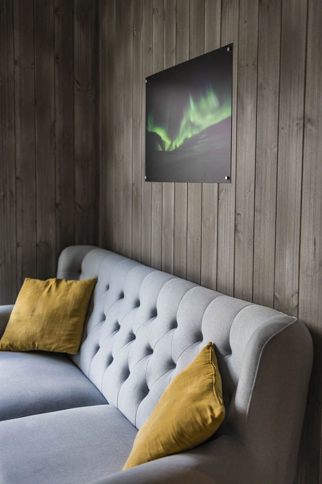 A light gray tufted sofa with yellow velvet pillows placed against a dark wooden paneled wall, featuring a framed photograph of the Northern Lights.