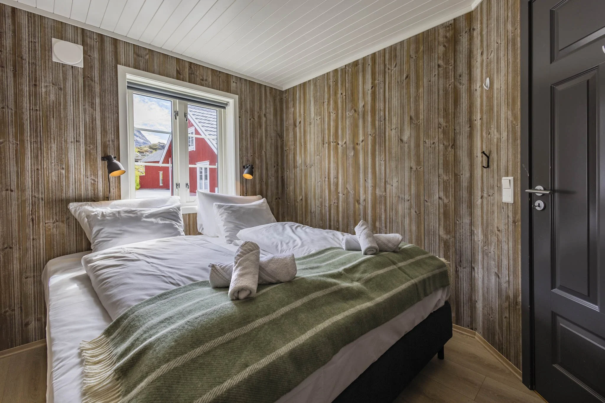 A bedroom with wooden wall paneling, a window showing a red house outside, a bed with white linens and two neatly folded towels on top, and black wall-mounted reading lights.
