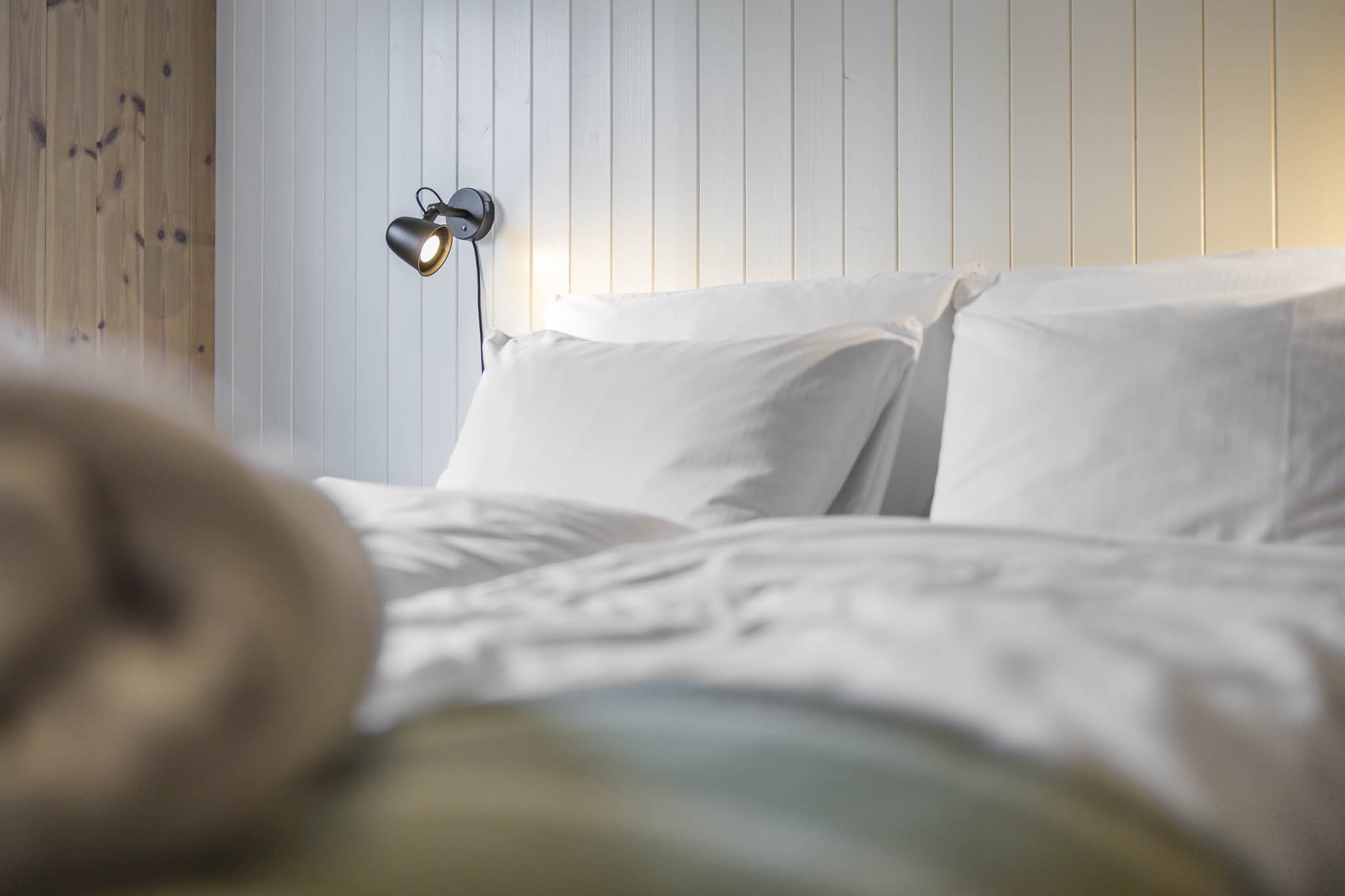 The image shows a bed with white sheets and pillows in front of a wooden wall with paneling. A black wall-mounted lamp is turned on above the bed. The photo is taken from a low angle, possibly from someone lying in bed.