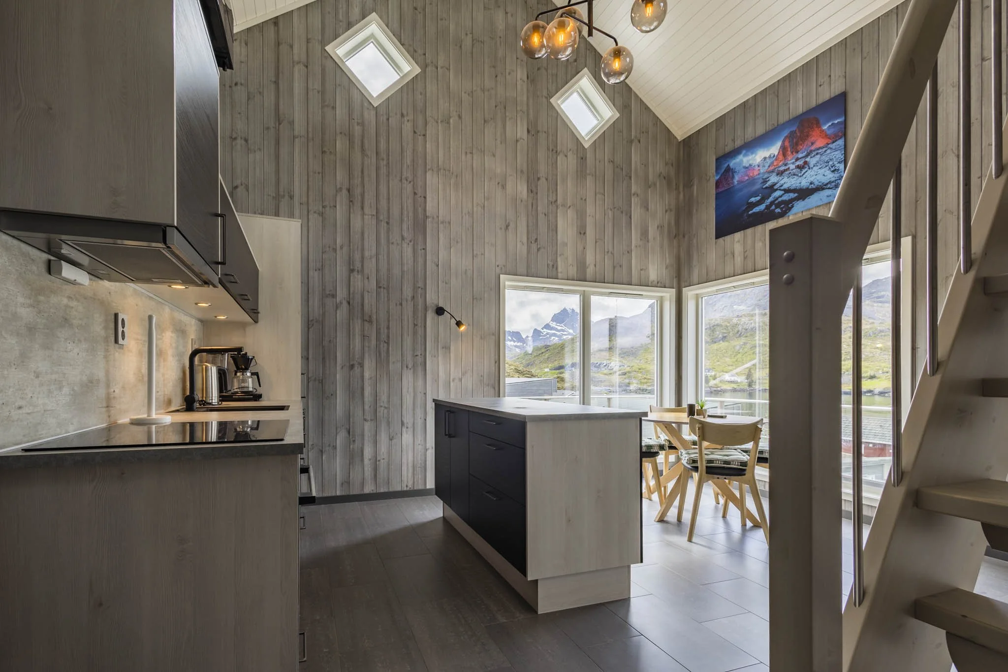 Interior view of a modern kitchen with wood-paneled walls, a kitchen island with a white countertop, and black lower cabinets. Large windows reveal a mountain landscape outside. A dining area with wooden chairs is visible near the windows. Skylights 