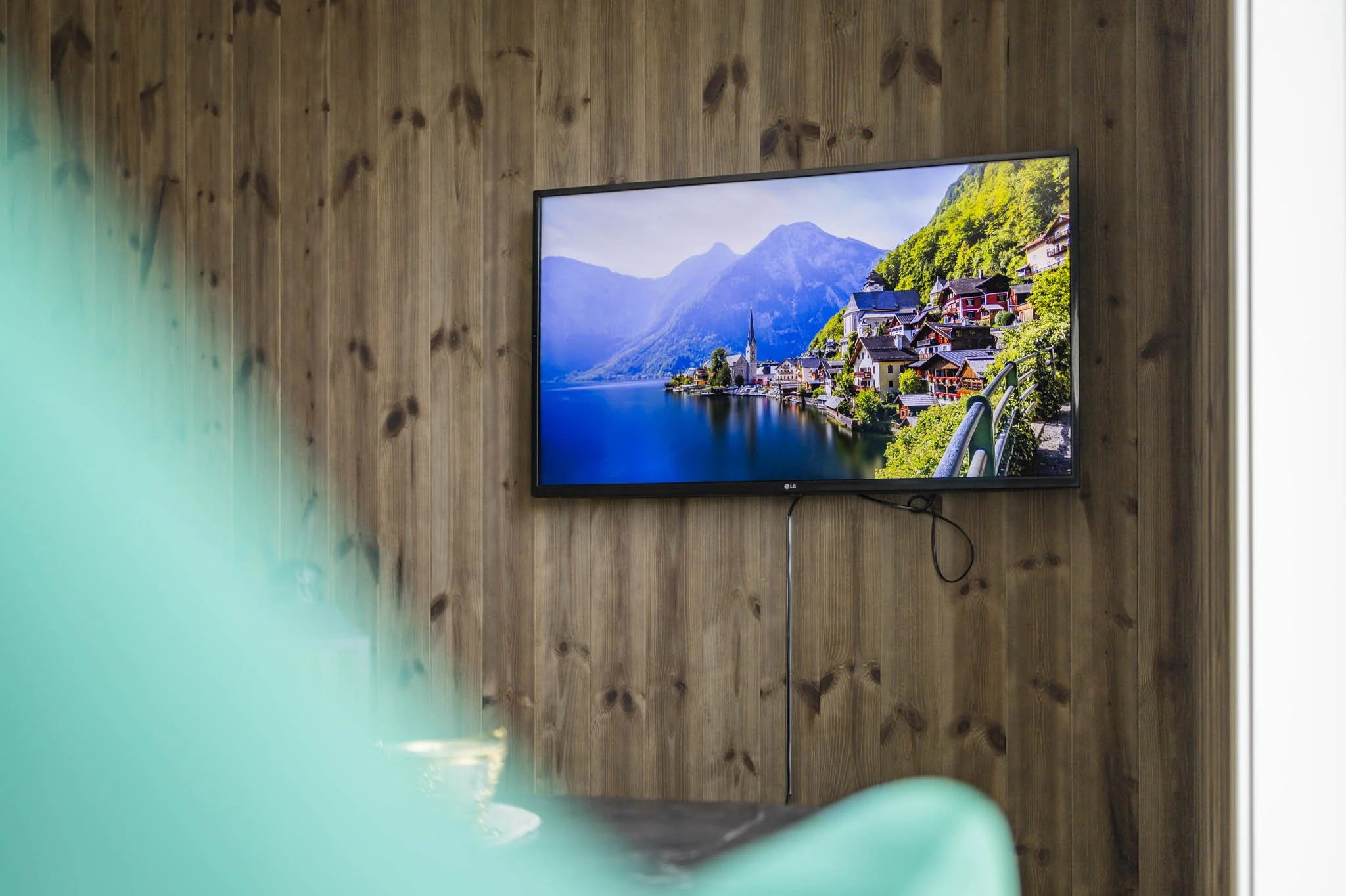 A flat-screen TV mounted on a wooden wall displaying a scenic view of a lakeside village with colorful houses, a church, and mountains in the background.