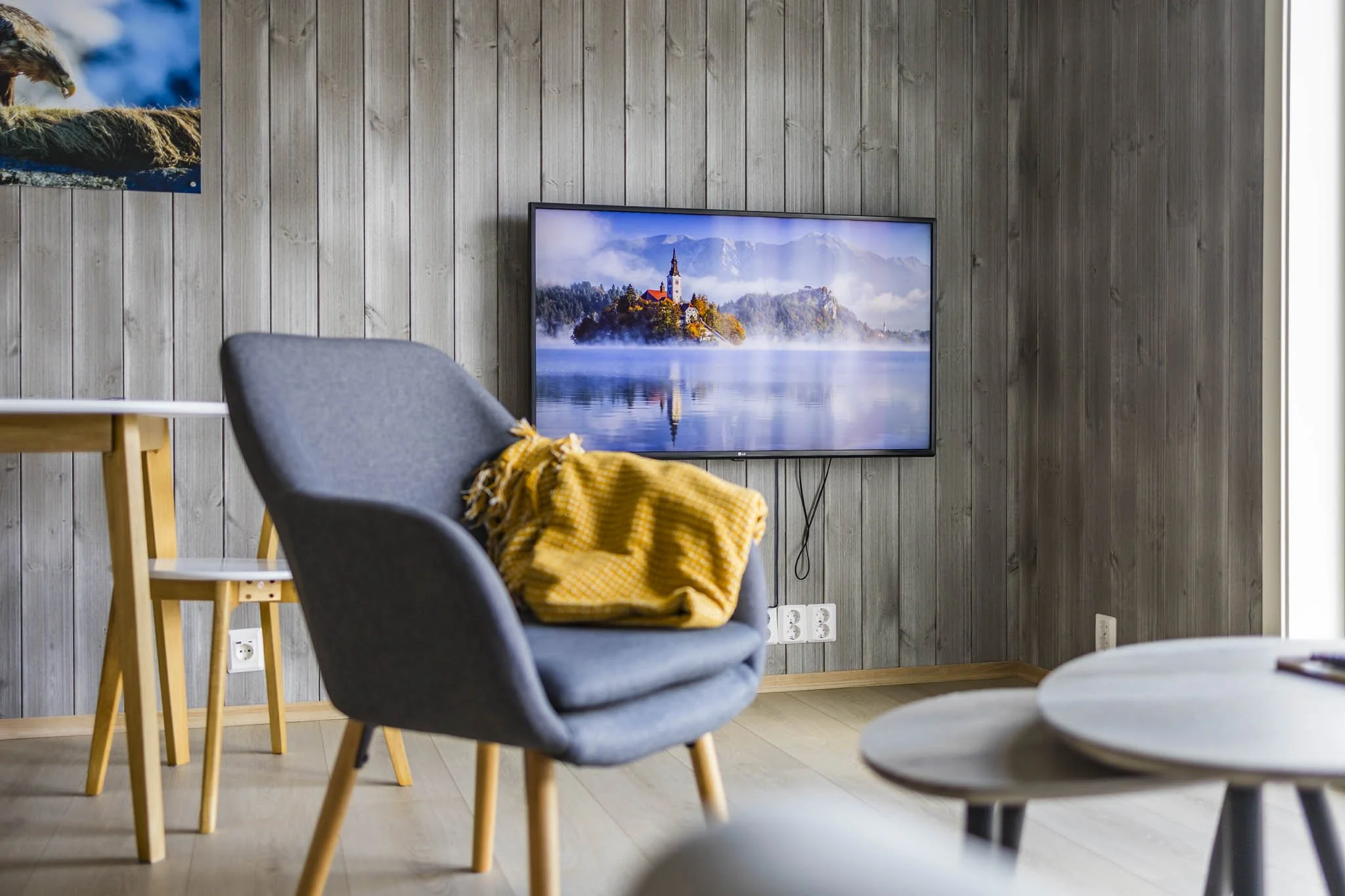 A living room with a flat-screen TV on the wall displaying a scenic image of a church on a small island surrounded by water and mountains in the background. There are gray chairs with wooden legs, and a yellow blanket draped over one of the chairs. T