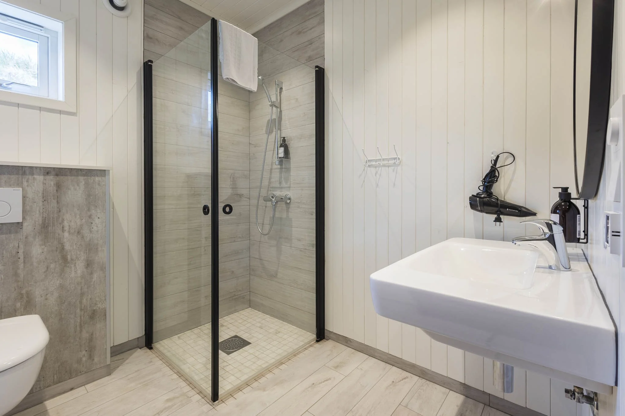Small bathroom with a corner shower with glass sliding doors, a wall-mounted shower head, a bottle of shampoo, a towel hanging outside the shower, a small window, a toilet, and a sink with a mirror above it.
