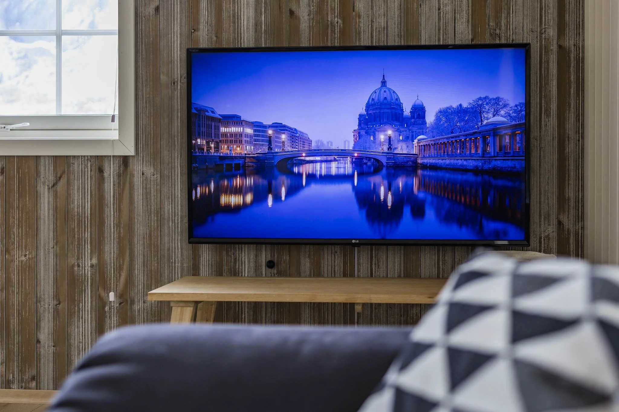 Living room with a large flat screen TV displaying a cityscape with a river and domed building, seen from a seating area with a striped pillow, wooden wall, and a window to the left.