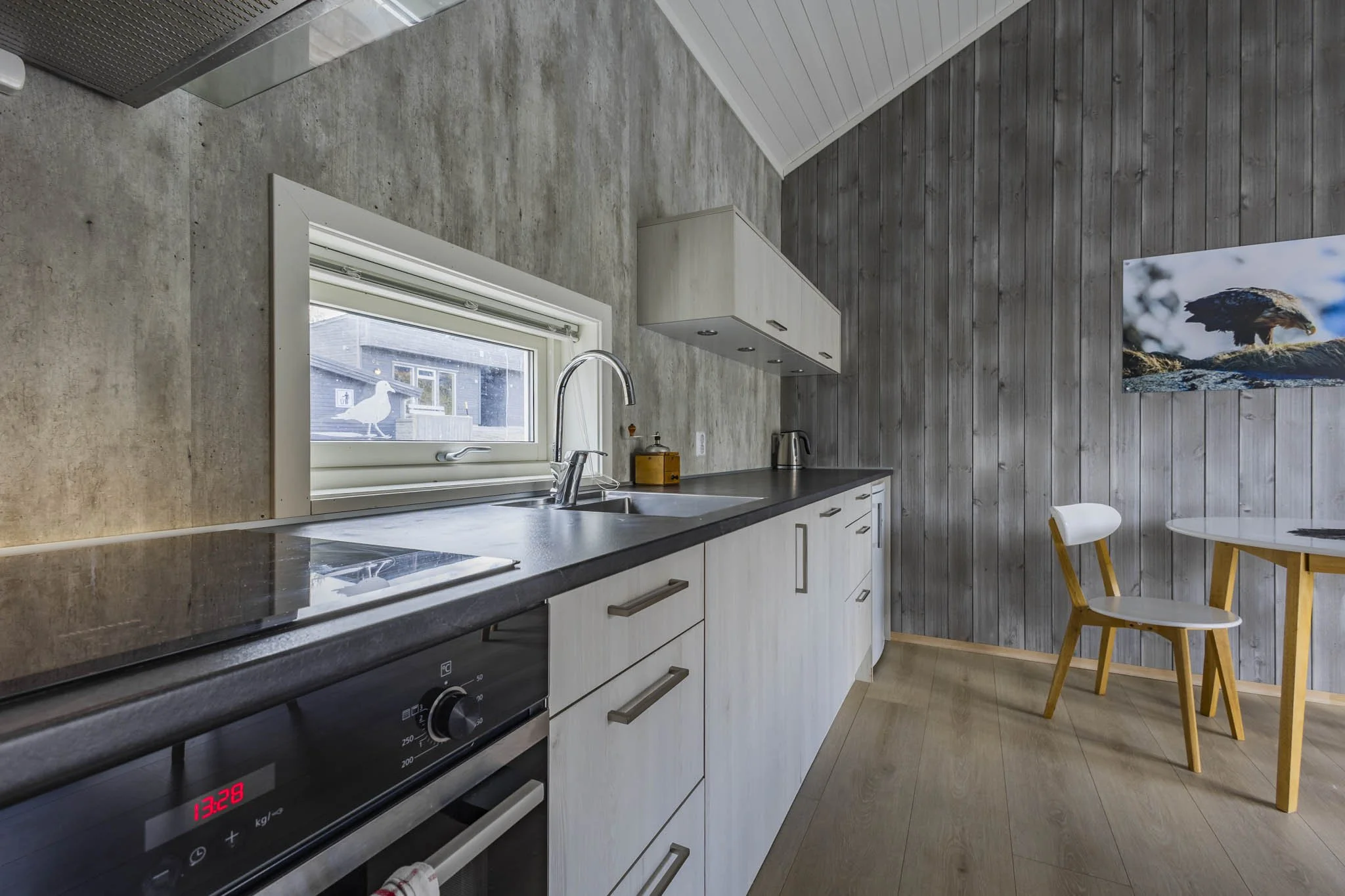 Modern kitchen with granite countertop, white cabinets, a window with a seagull outside, wooden dining chairs, and a wall with wood paneling and a mountain eagle photograph.