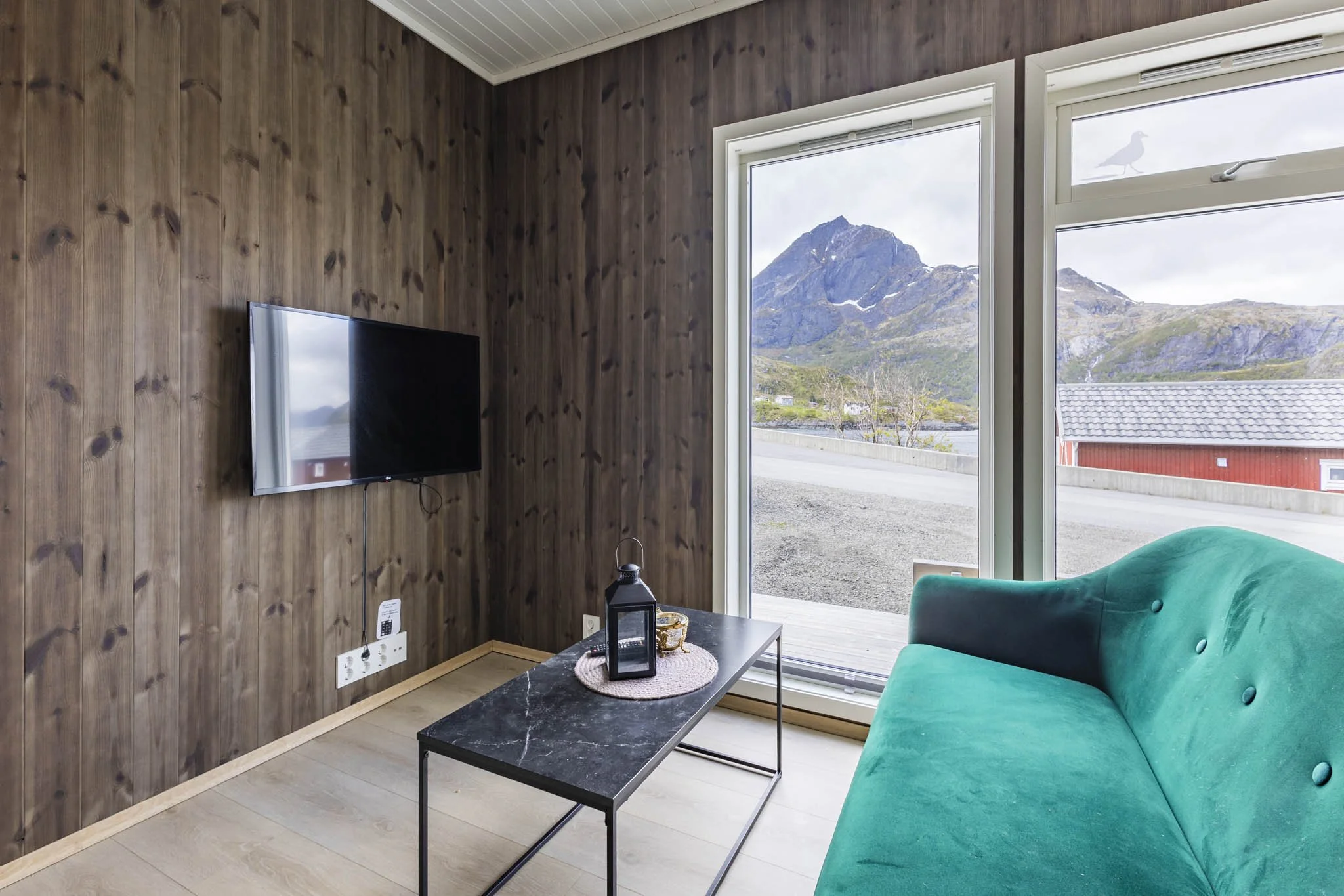 Living room with wood-paneled walls, a mounted flat-screen TV, large windows showing mountain scenery, a teal sofa, and a black marble coffee table.
