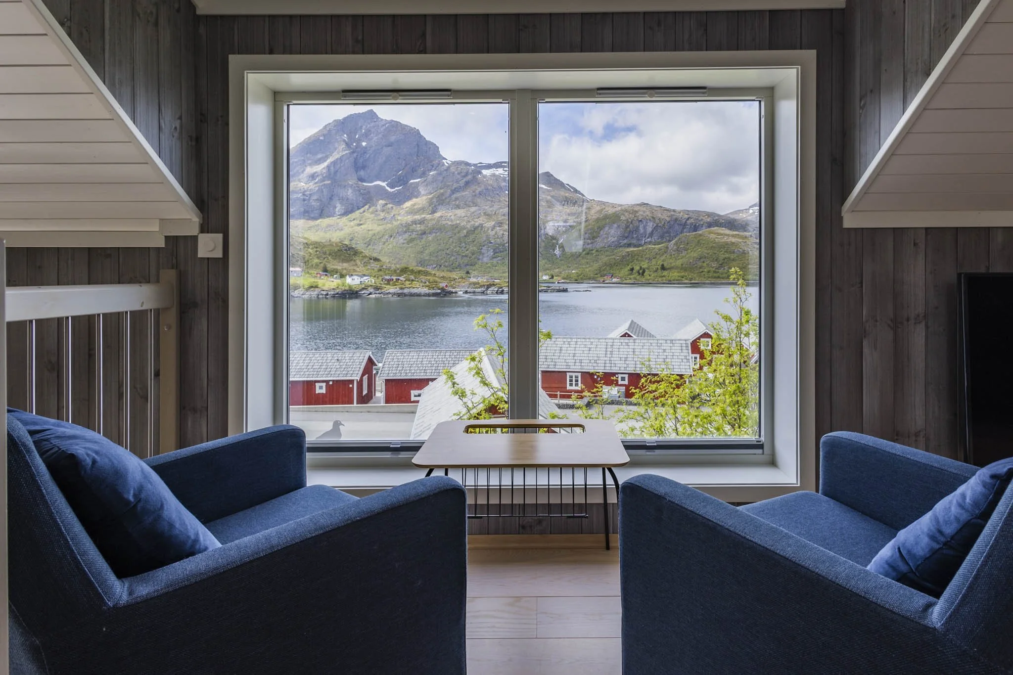 Room with a large window showing mountains, water, and a cloudy sky outside, with dark wood wall paneling inside.