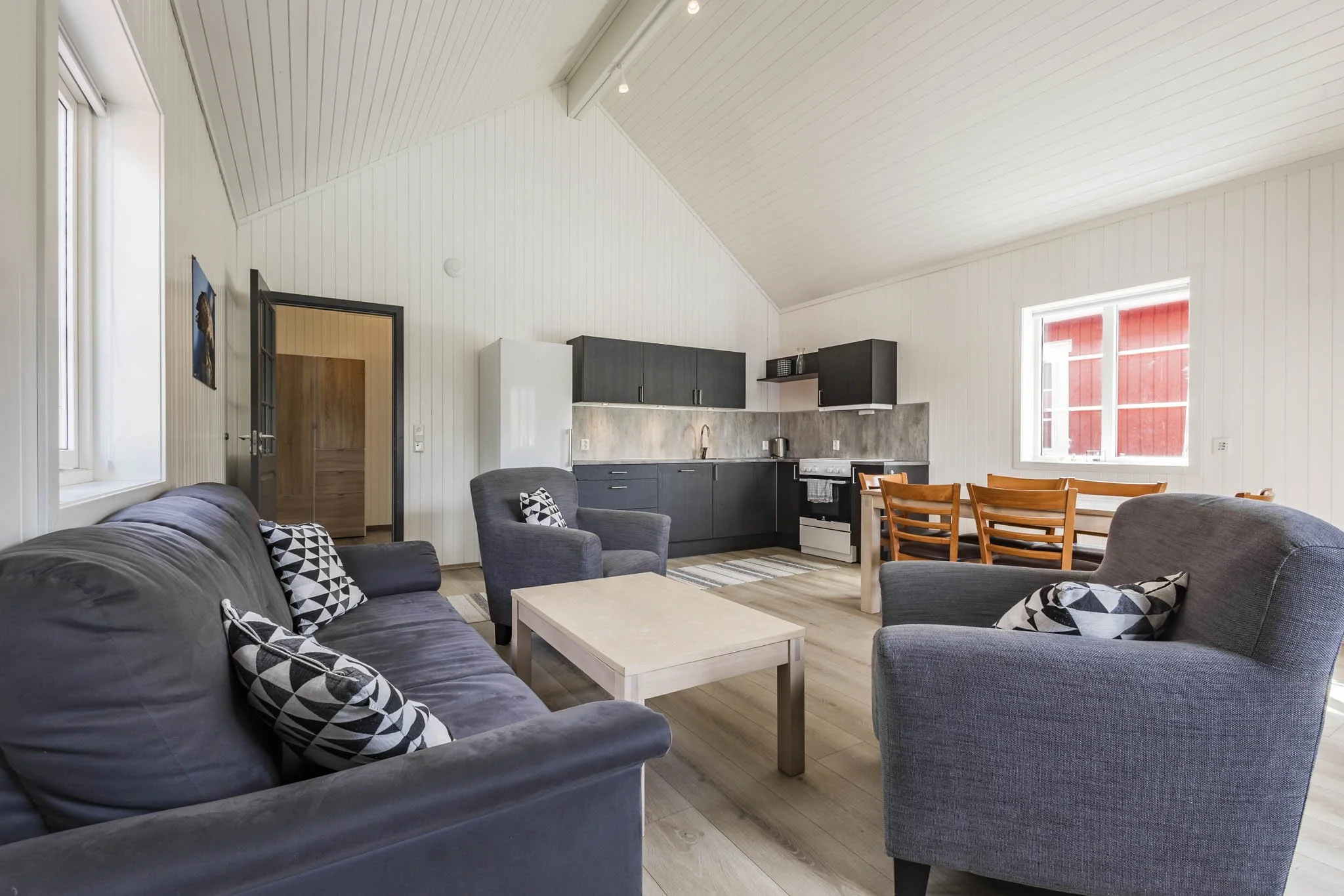 A living room with a dark gray sofa and matching armchair, a light-colored coffee table, and patterned throw pillows. Behind the sofa is a wall with a window, and nearby are some chairs and a dining table, with the kitchen area featuring black cabine