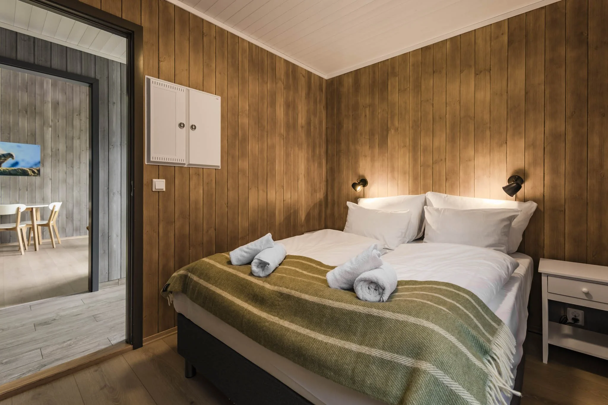 A cozy bedroom with wooden paneled walls and white linens on the bed. Towels are rolled and placed at the foot of the bed. A small white nightstand and reading lights are mounted on the wall above the bed. An open doorway leads to a dining area with 