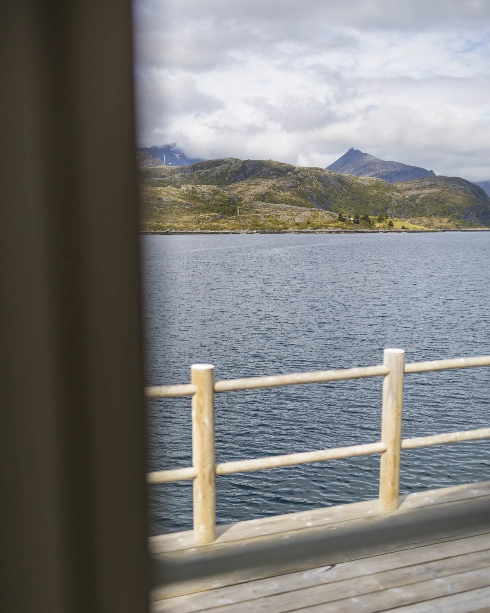 View of mountain landscape with a lake in the foreground, seen through a window with curtains and a railing.