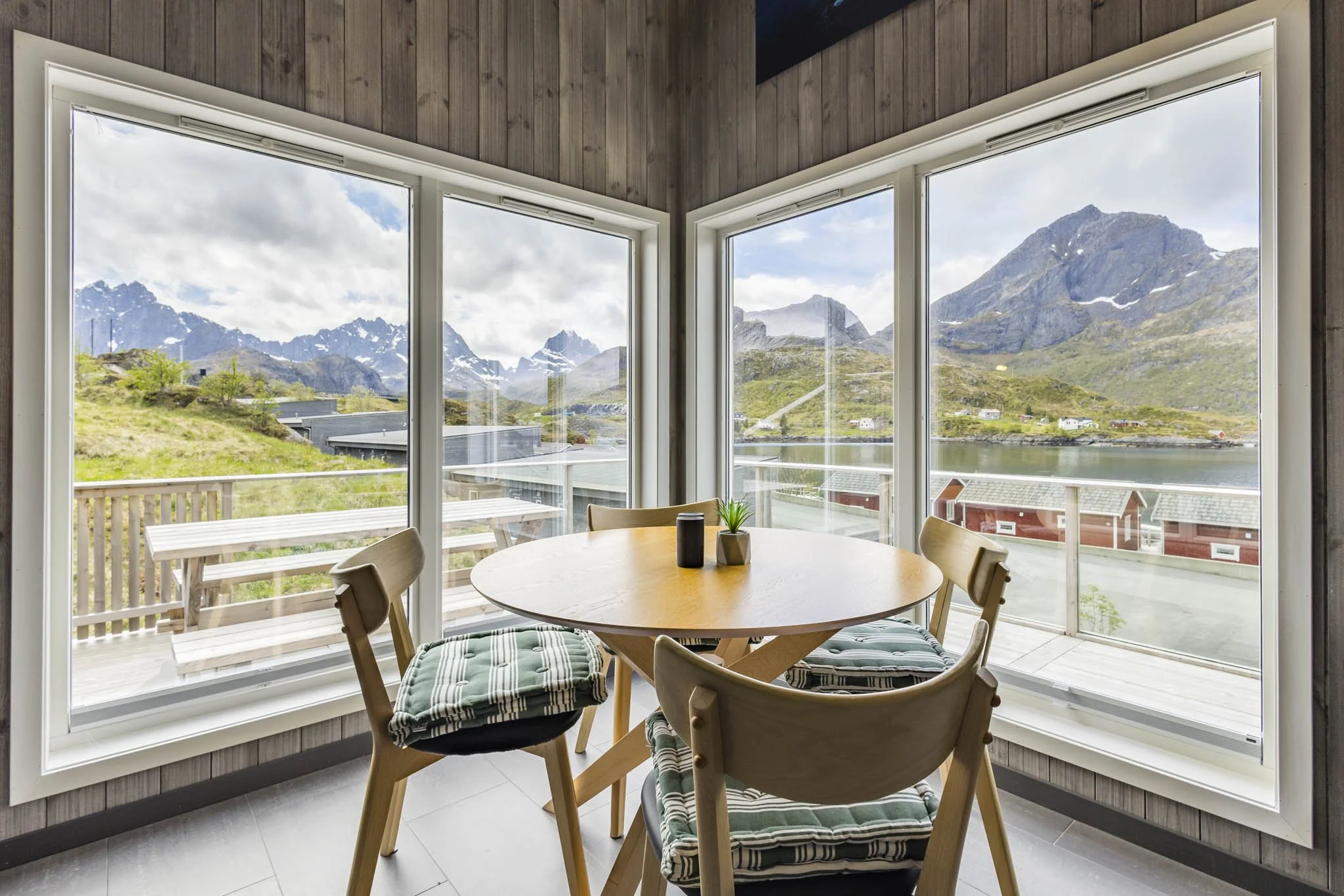 View through large floor-to-ceiling windows showing mountains, a lake, and a small town with houses, taken from inside a wooden-paneled room with a round dining table and chairs.