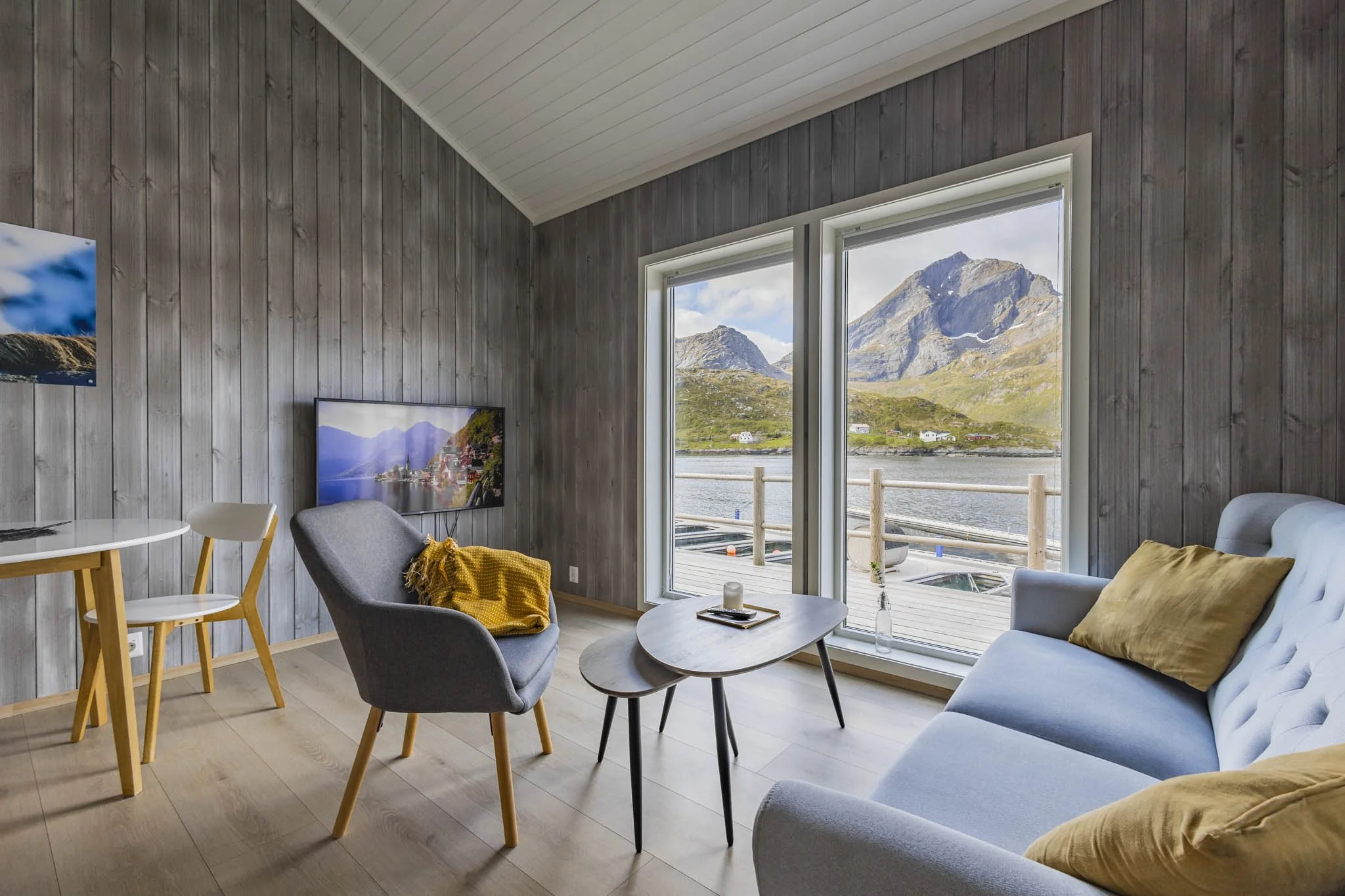 Living room with wood-paneled walls and large windows showing a mountain landscape and water. Contains a gray armchair with a yellow blanket, a round coffee table with a bottle and remote, a light blue sofa with yellow cushions, a wall-mounted TV displaying a mountain scene, and a dining table with white chairs.