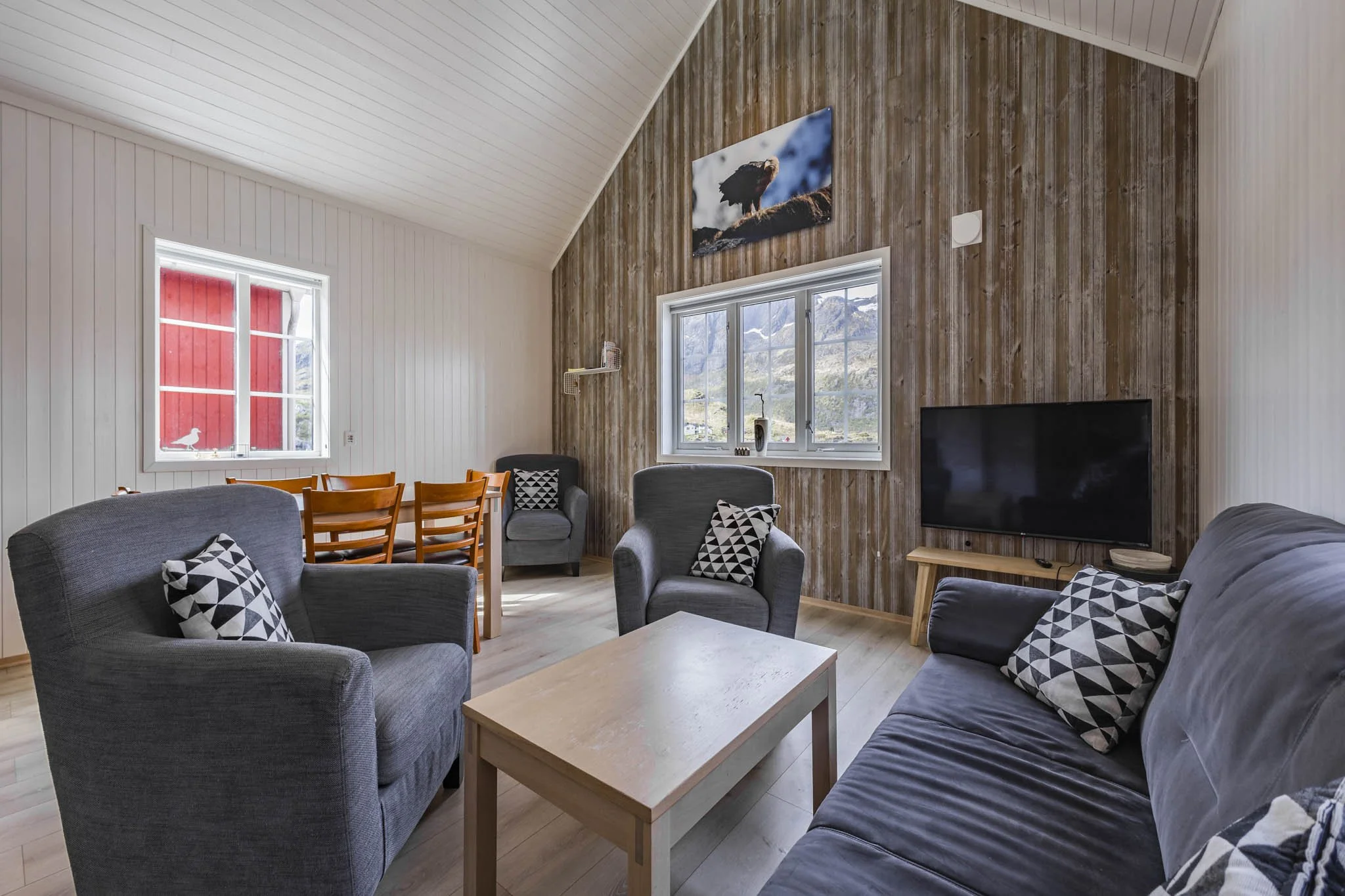 Living room with gray sofas, black and white geometric pillows, wooden coffee table, large window with mountain view, wooden wall with mounted photograph of an eagle, flat-screen TV, and striped white and natural wood ceiling.