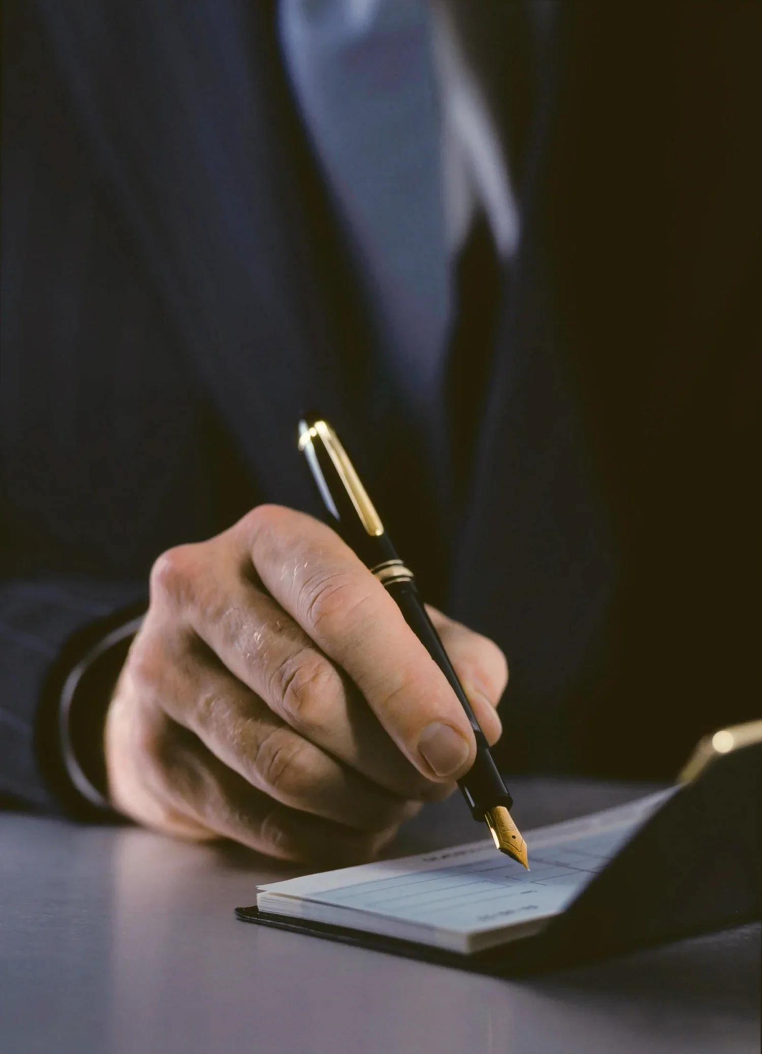 A person dressed in a dark suit writing in a notepad with a gold fountain pen.