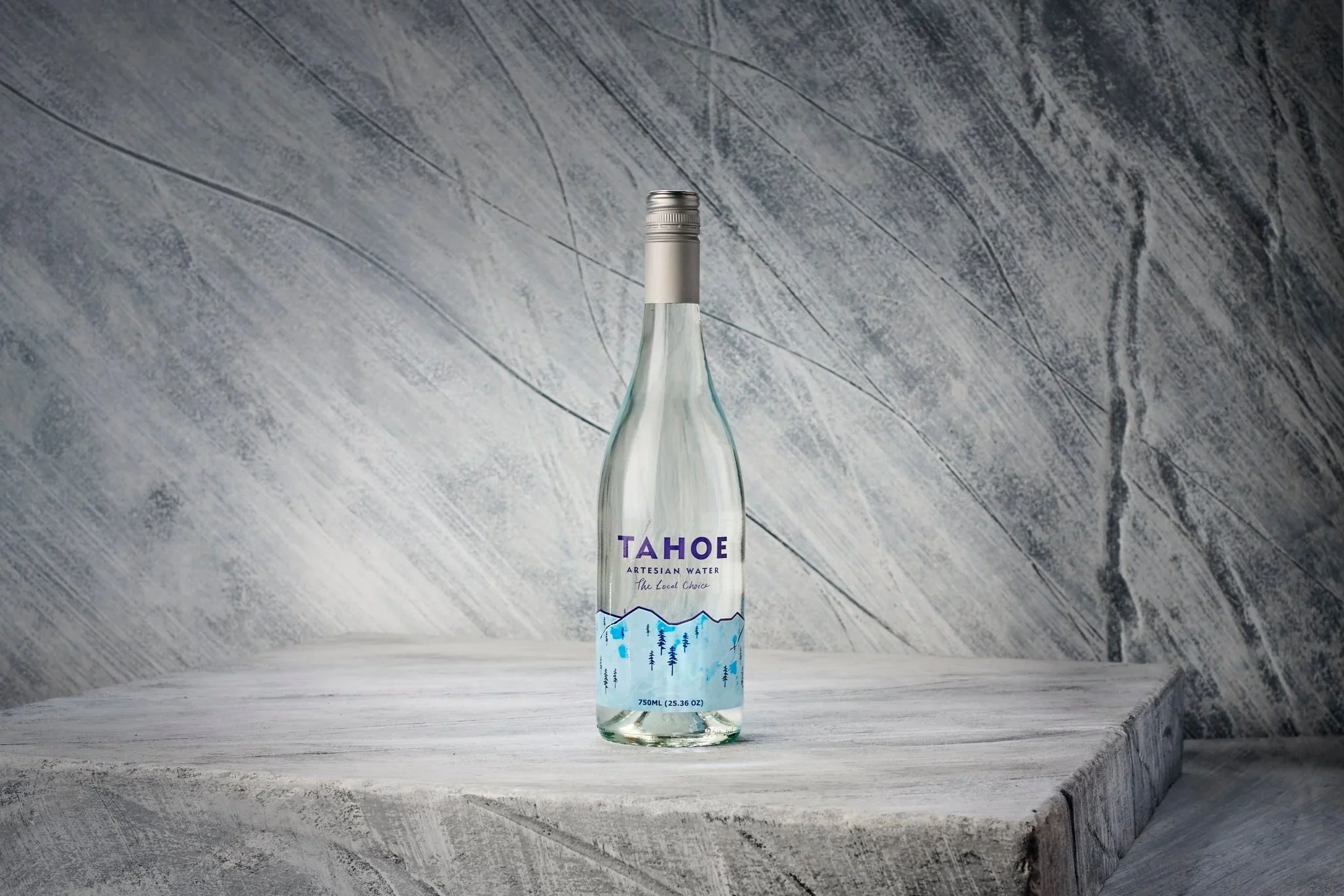 A clear glass bottle of Tahoe Artesian Water with a silver cap, standing on a textured white wooden surface against a gray marbled background.