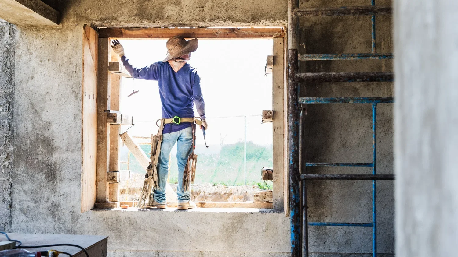 Construction worker standing at a large window opening inside a building, facing outside, wearing a hat and safety tools.