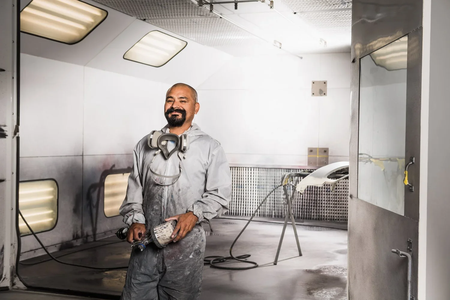 A man in a gray work jumpsuit holding a spray gun, standing in a spray booth with bright lighting, white walls, and an industrial setting.