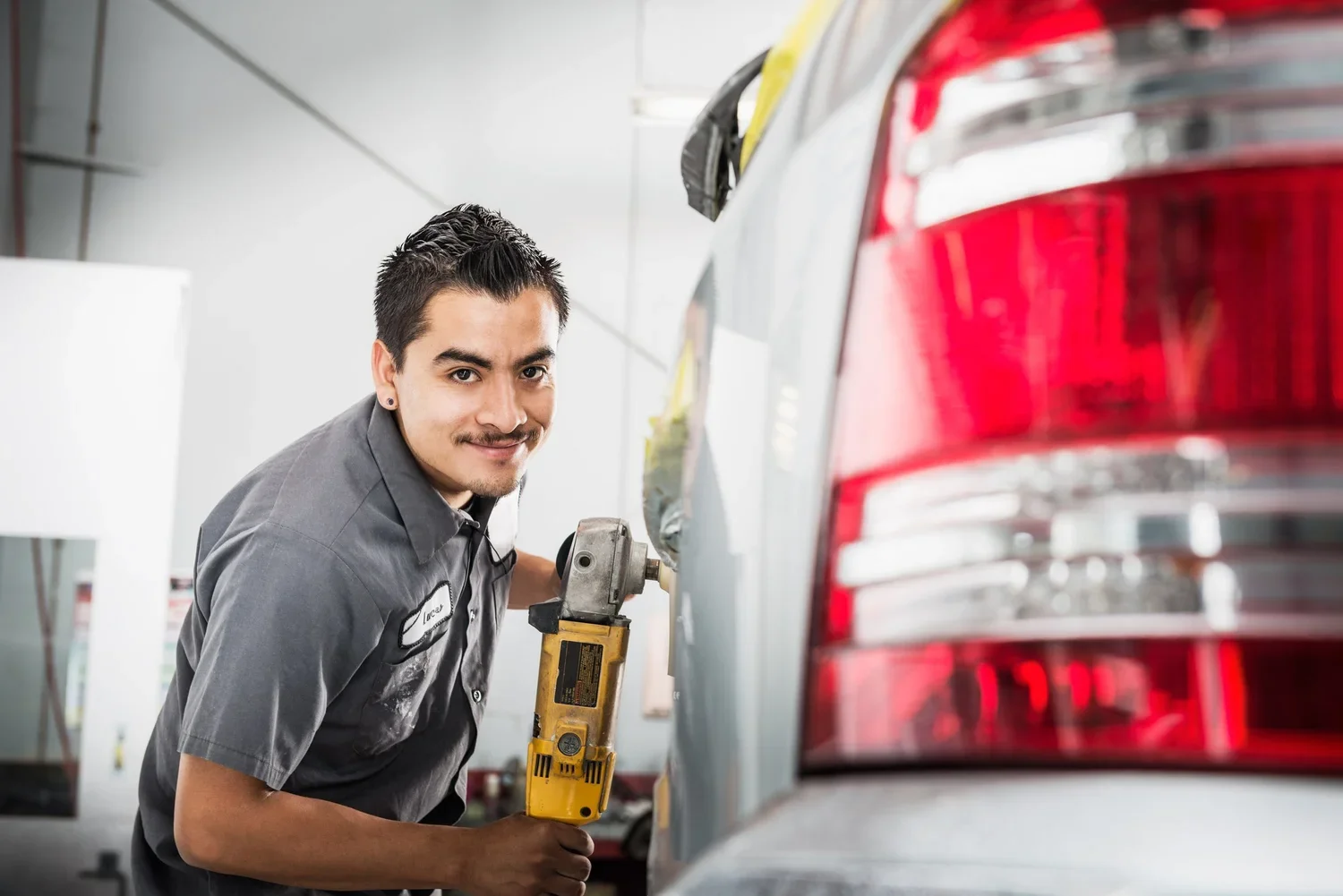 A mechanic at an auto repair shop using a power tool on the back of a silver SUV, smiling at the camera.