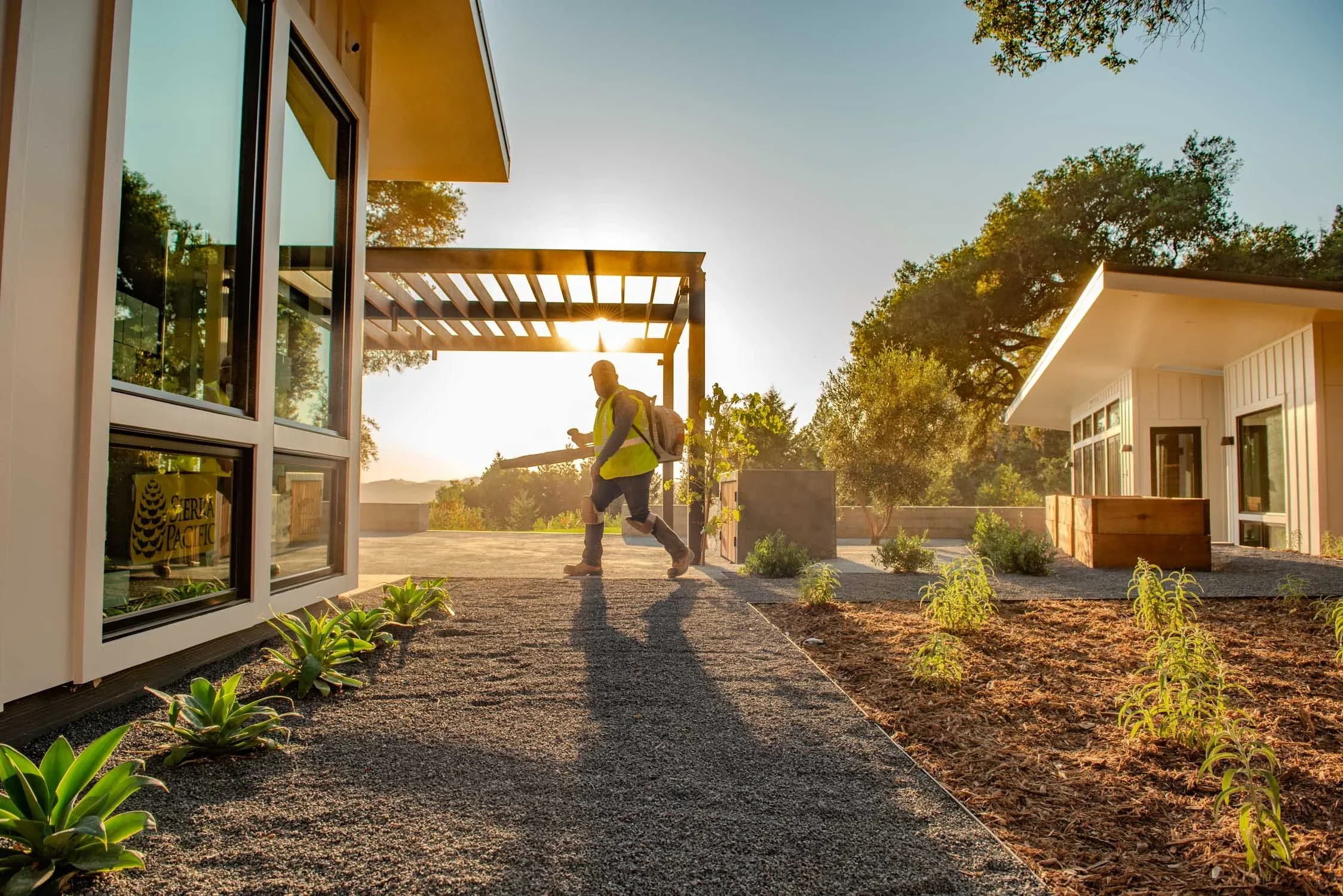 Person walking on a gravel path in a modern residential neighborhood during sunset, carrying a backpack.