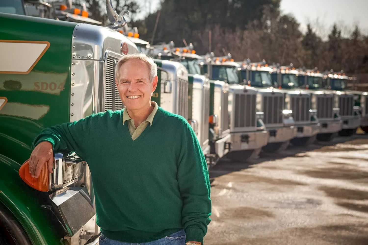 A smiling man in a green sweater standing next to a row of green and silver semi-trucks outdoors.