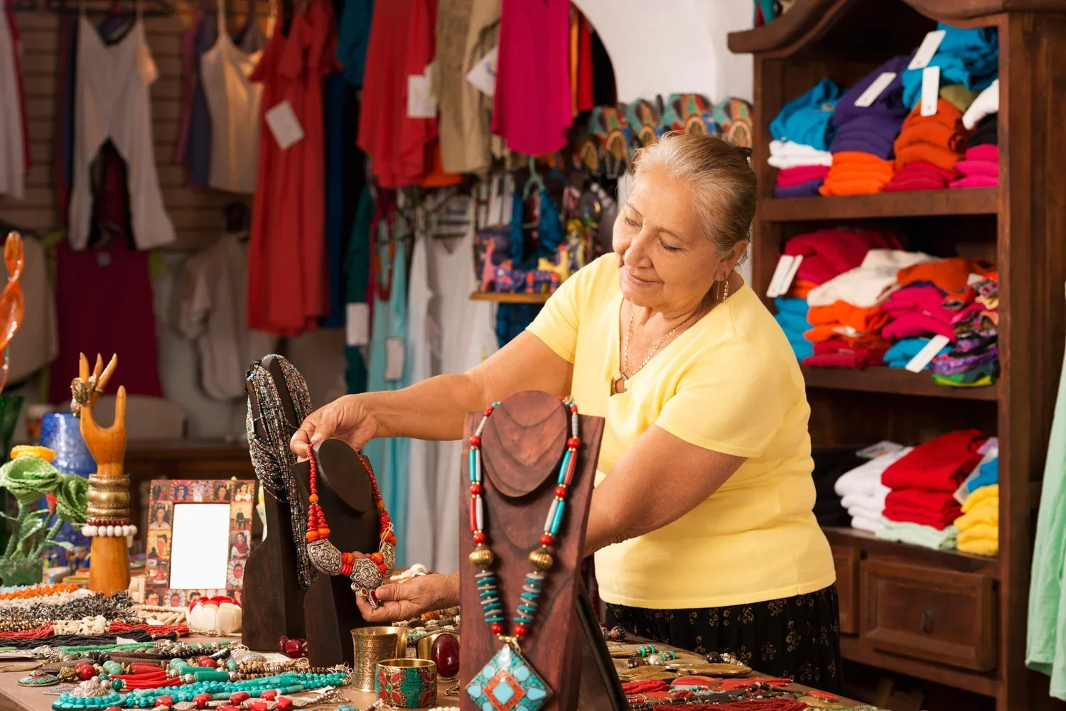 A woman shopping for jewelry at a store with colorful clothing and accessories displayed around her.