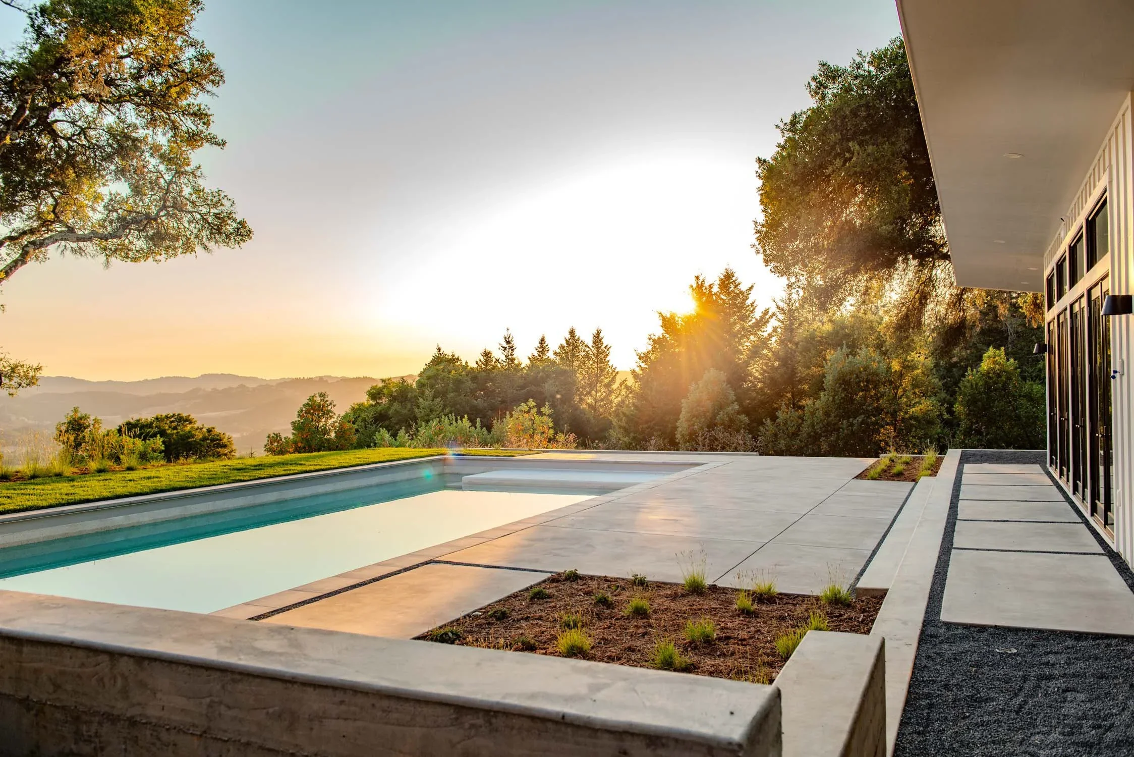 Modern backyard with a swimming pool, concrete patio, and garden beds, overlooking scenic hills at sunset.