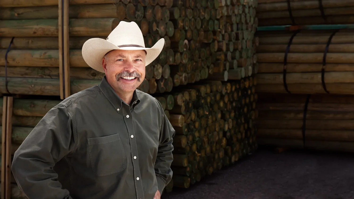 A man wearing a cowboy hat and dark button-up shirt stands in front of stacked lumber at a lumber yard.