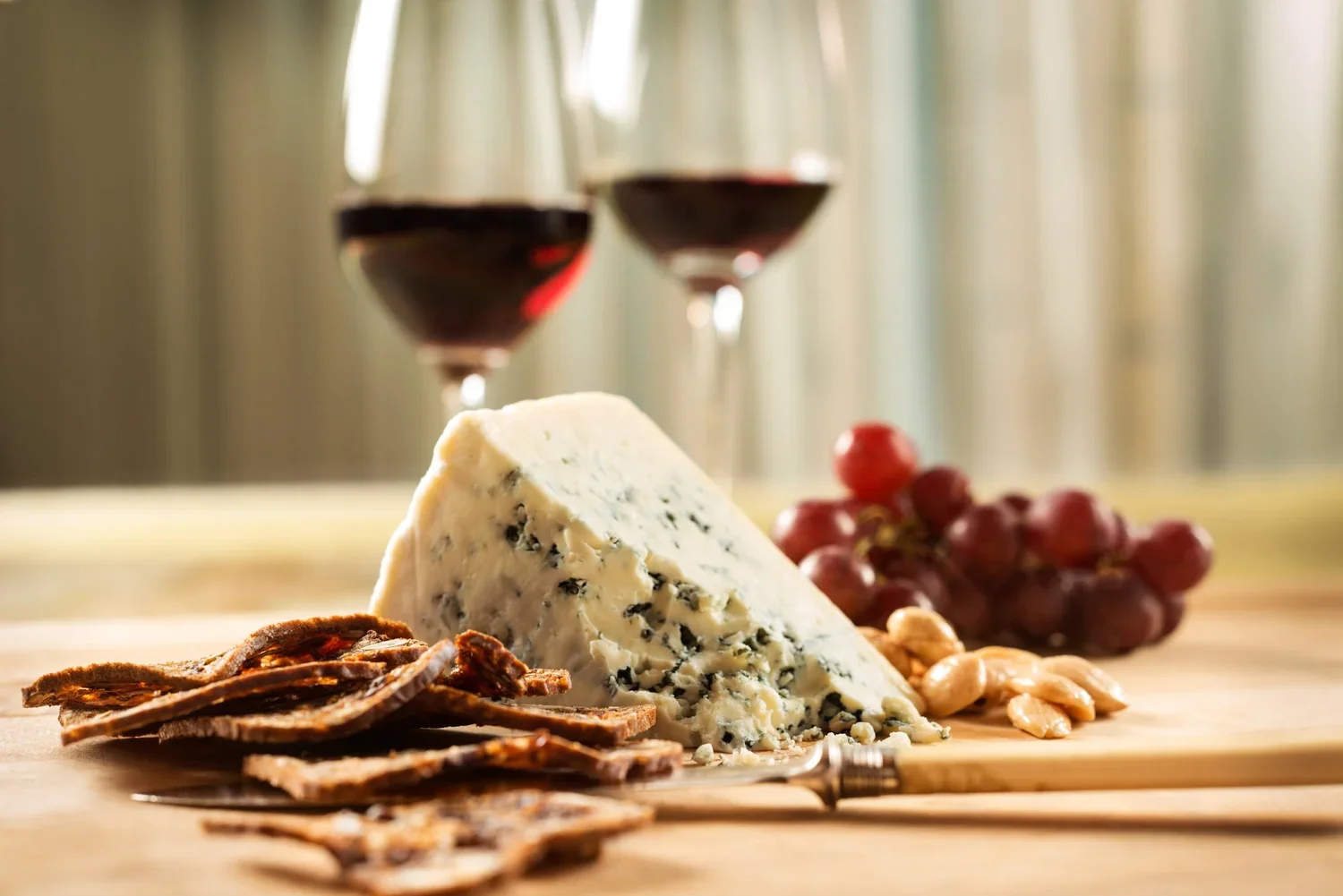 A wedge of blue cheese on a wooden surface, surrounded by dried meats, a bunch of red grapes, and cashew nuts, with two glasses of red wine in the background.